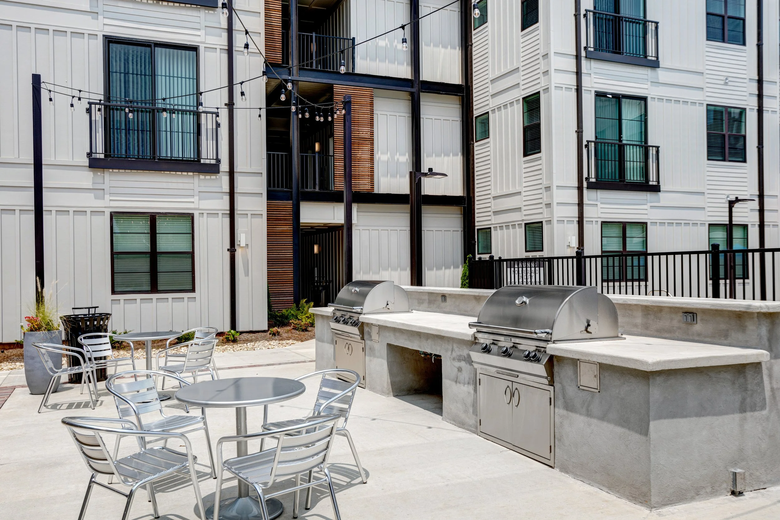 An outdoor patio area with metal tables and chairs, built-in outdoor grills, string lights, and a modern apartment building with balconies in the background.
