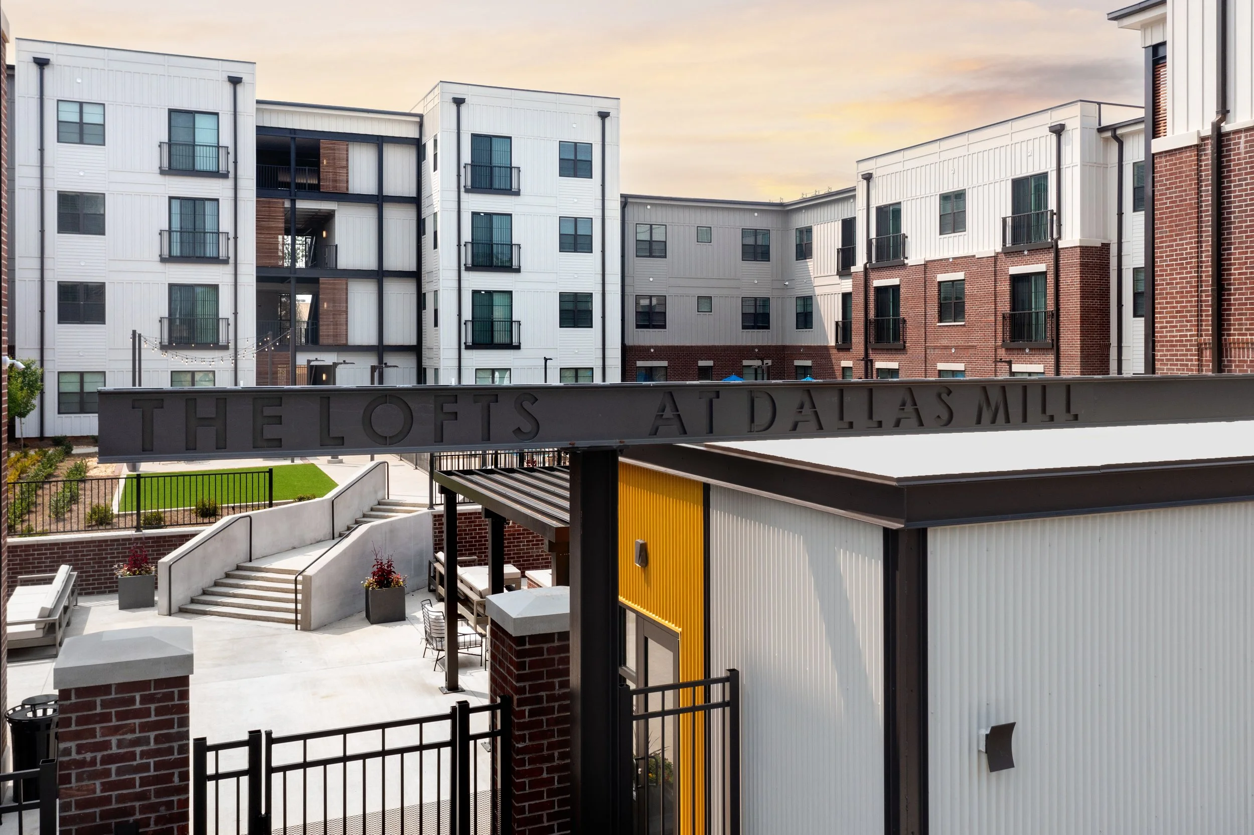 Modern apartment complex with white and red brick facades, balconies, and a courtyard with stairs and string lights, at sunset.