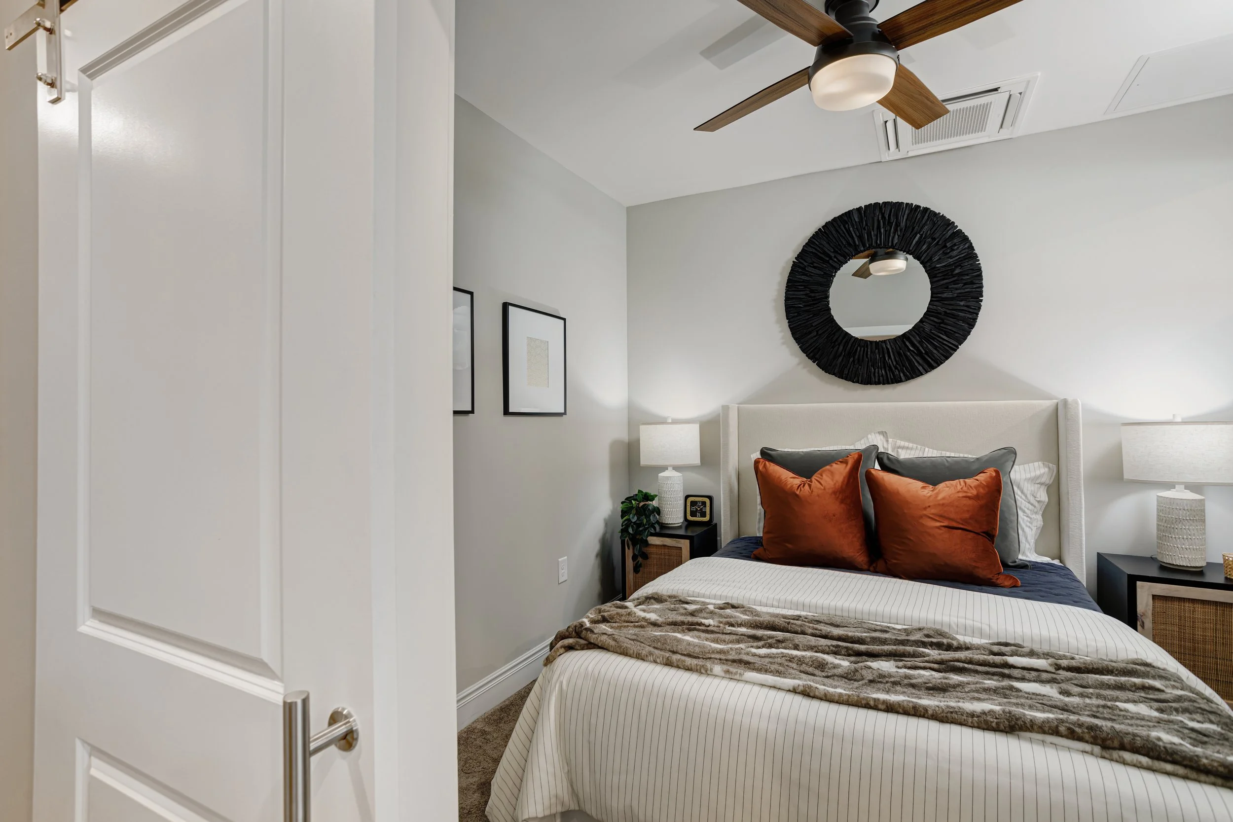 A bedroom with a beige bed, dark blue and rust-colored pillows, a fur throw blanket, and white striped bedding. There are two nightstands with table lamps, a potted plant, and a digital clock. The wall above the bed has a round black decorative mirro