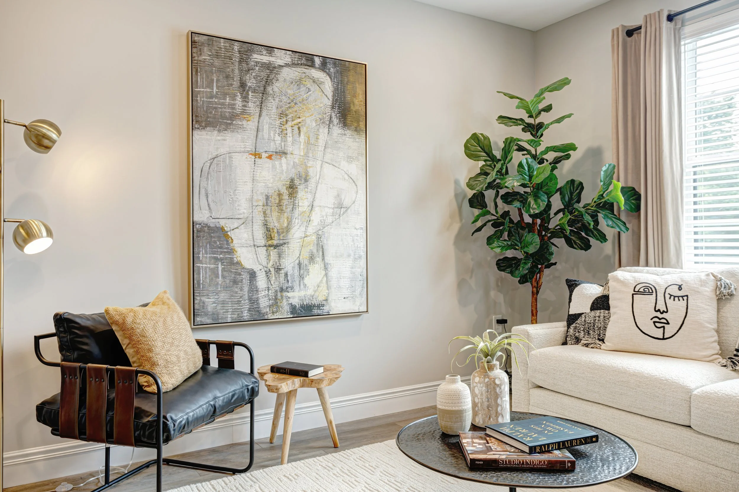 A cozy living room with beige walls, a large abstract art piece, a beige sofa with decorative pillows, a black armchair with a pillow, a wooden side table, a small round coffee table with books, a potted plant, and a window with striped curtains.