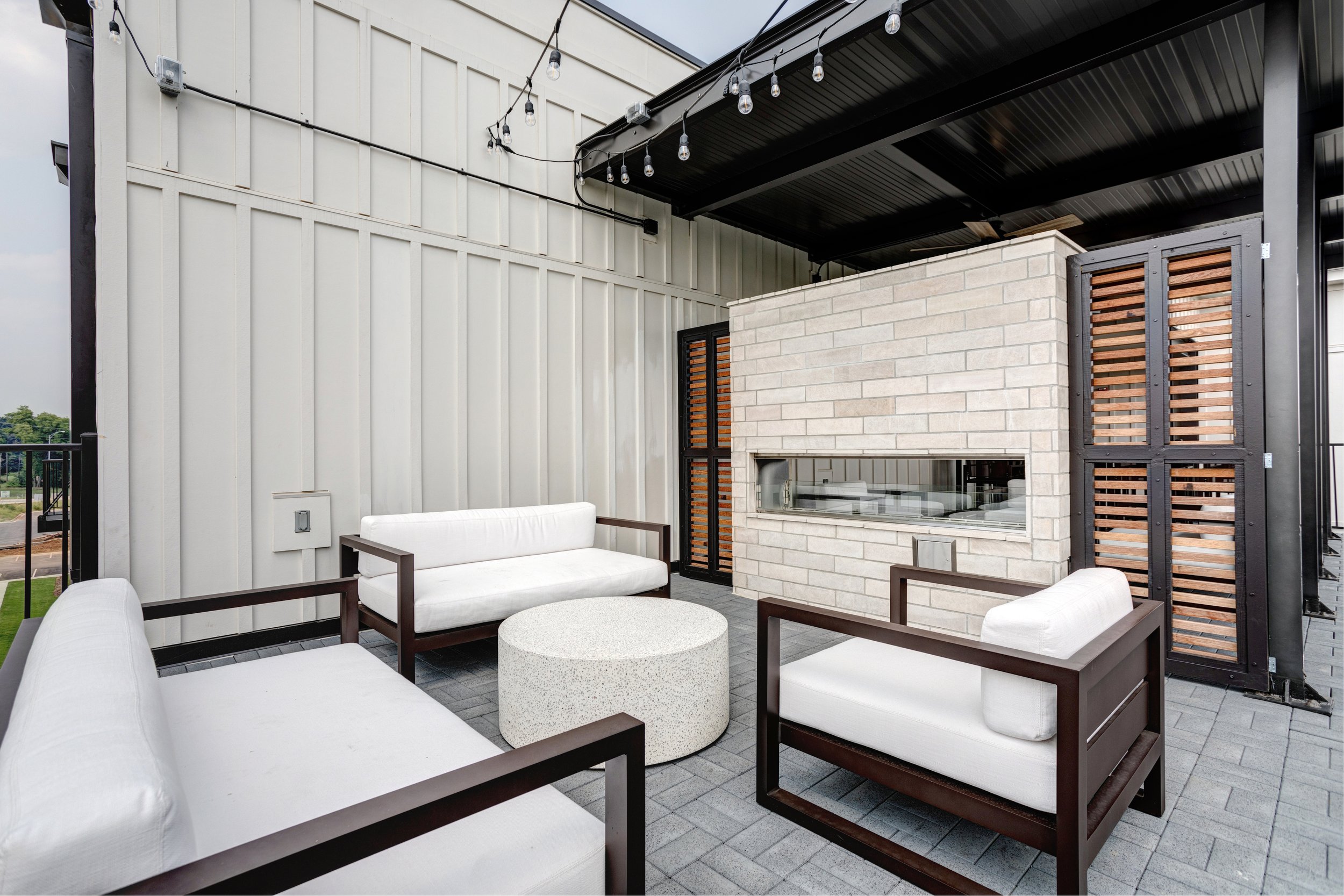 Outdoor patio with white cushioned seating, a round coffee table, a brick fireplace, and string lights hanging from the black ceiling. Modern design with a mix of metal, brick, and wood materials.