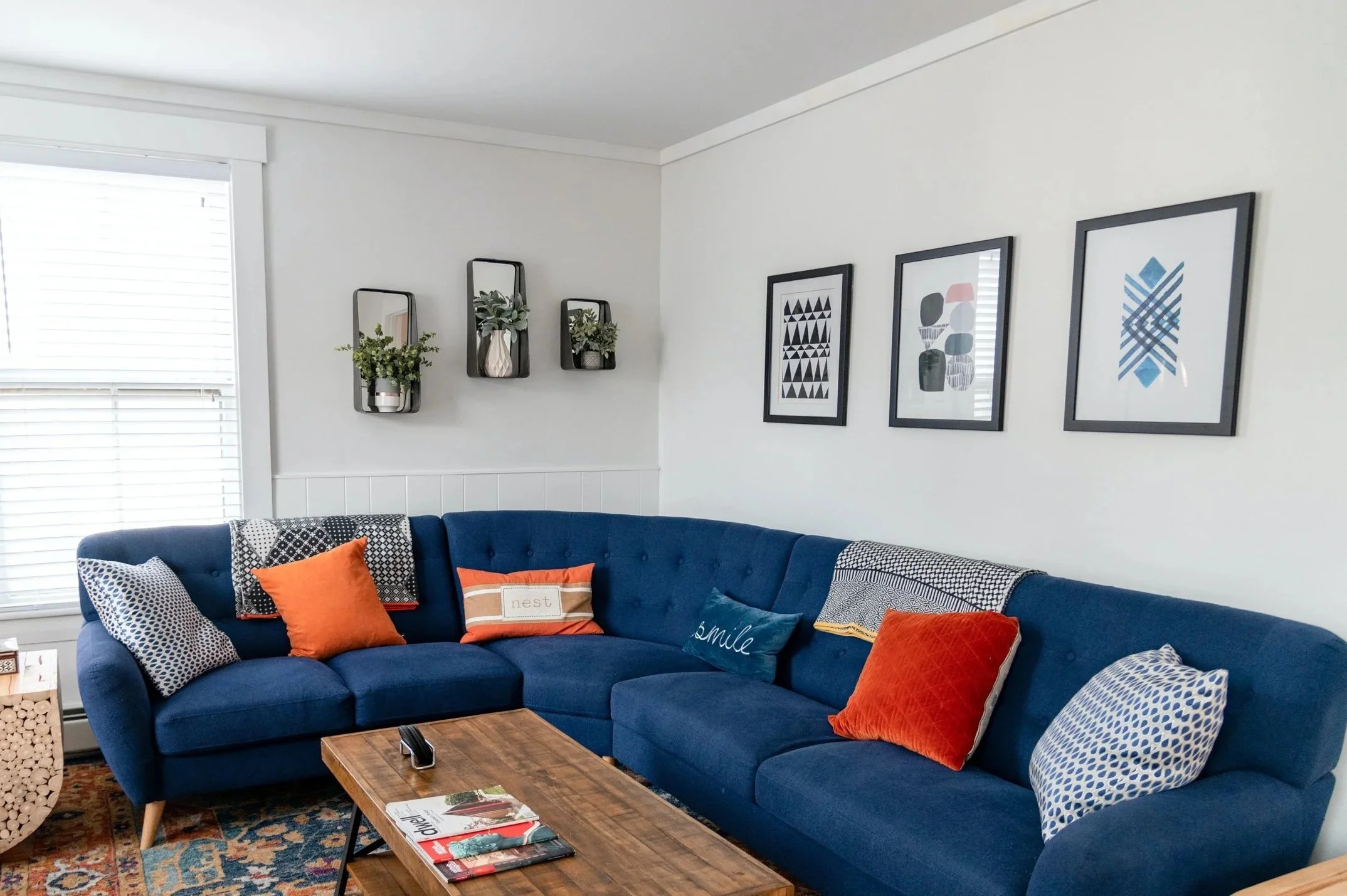 Living room with a blue sofa decorated with orange and patterned pillows, a wooden coffee table, framed abstract art on the walls, and windows with white blinds.