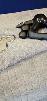 image of percussion massage gun and massage scraping tool sitting on a grey towel on a massage bed