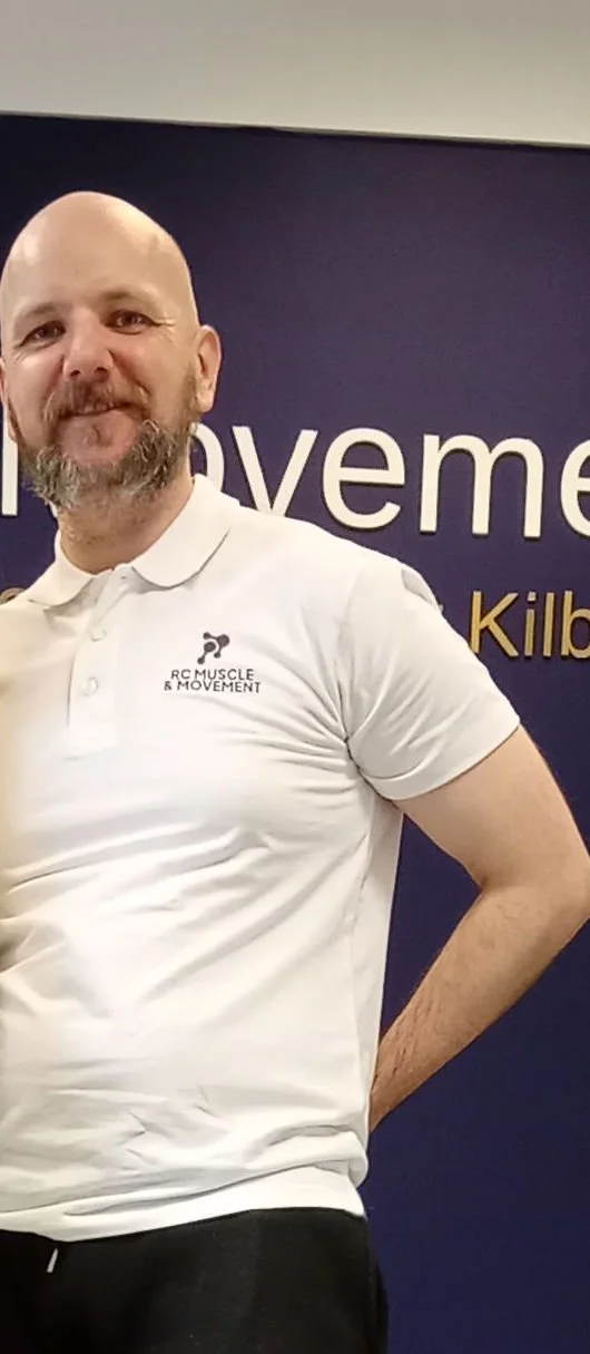 A man with a beard and bald head wearing a white polo shirt with a logo that says 'RC MUSCLE & MOVEMENT,' standing in front of a dark blue background with partial white and gold text.