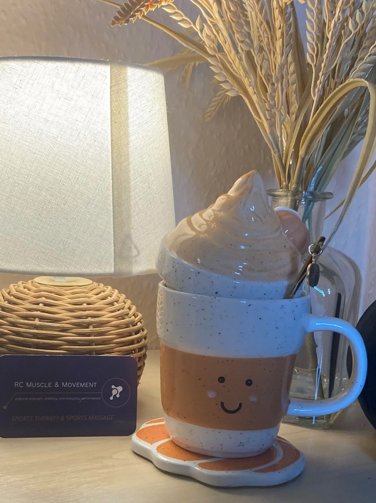 A beige ceramic mug with a smiling face design, filled with whipped cream or foam, placed on a saucer, with a small smiley face painted on it. Behind the mug, there is a clear glass jar with dried wheat or grass stems. To the left, there is a woven rattan box and a white textured lampshade.