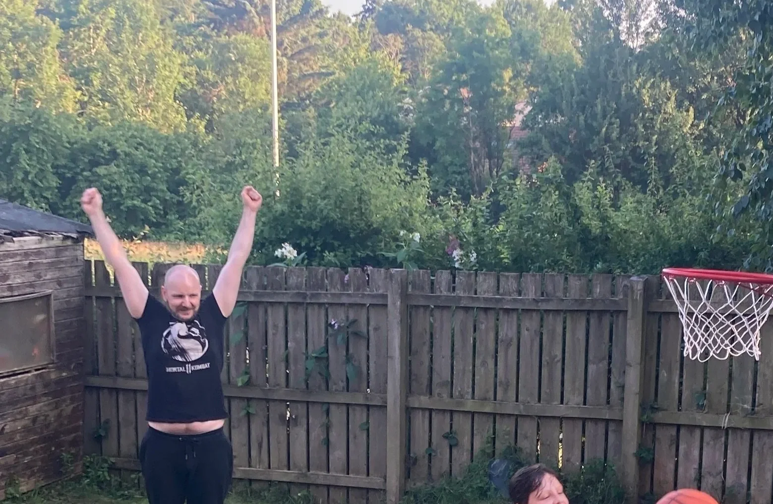 A man with a bald head and beard raising his arms in celebration outside, wearing a black T-shirt with a Mortal Kombat logo, next to a wooden fence and a basketball hoop.