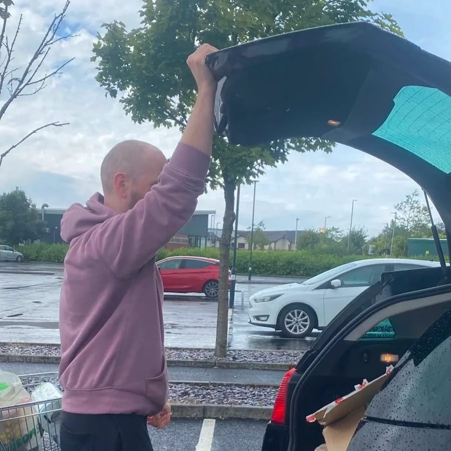 A person with a bald head and light purple hoodie loading groceries into the trunk of a black car in a parking lot on a rainy day.