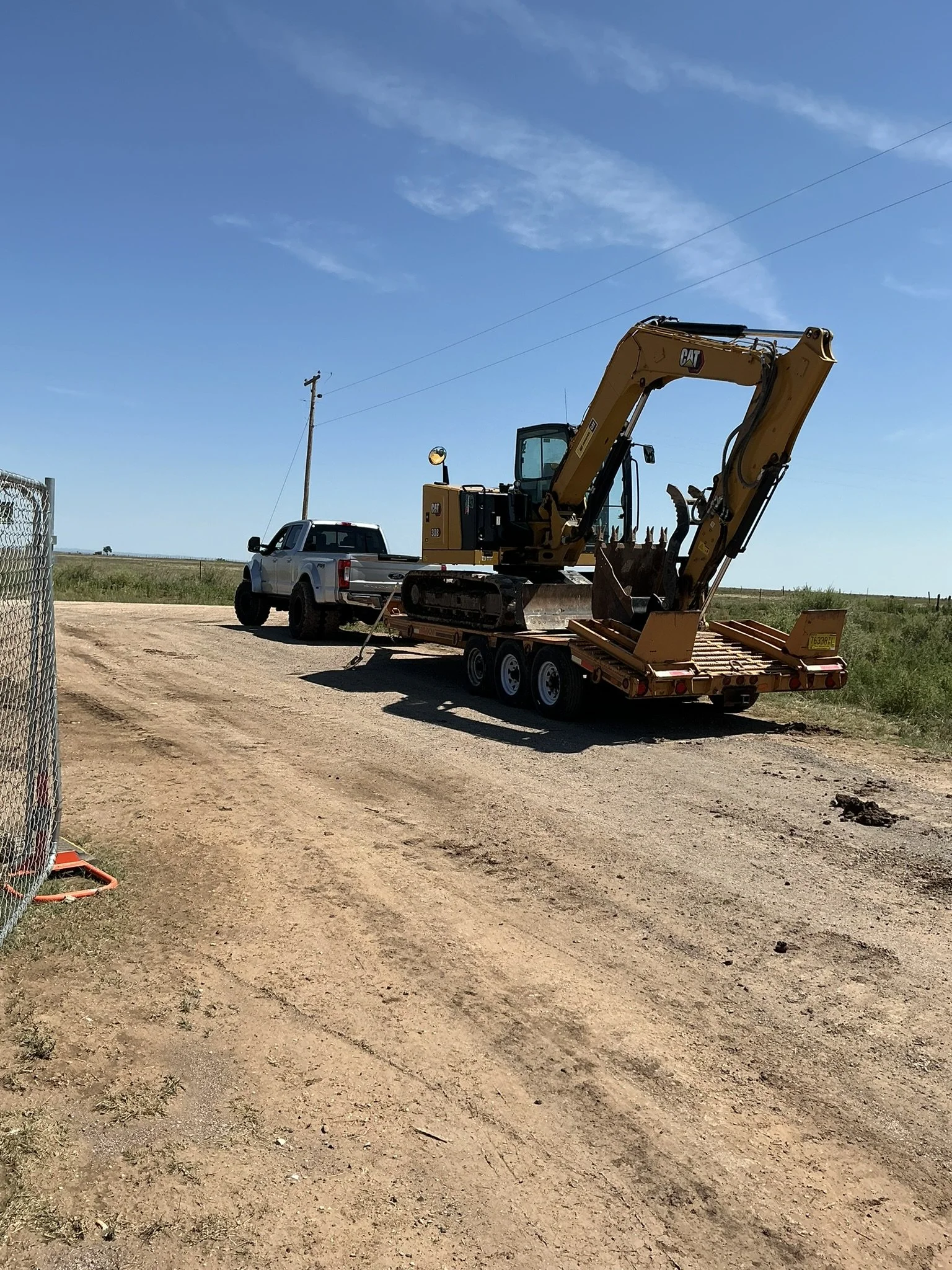 A yellow Caterpillar excavator on a trailer hitched to a pickup truck on a dirt road in an open field with a utility pole and blue sky.