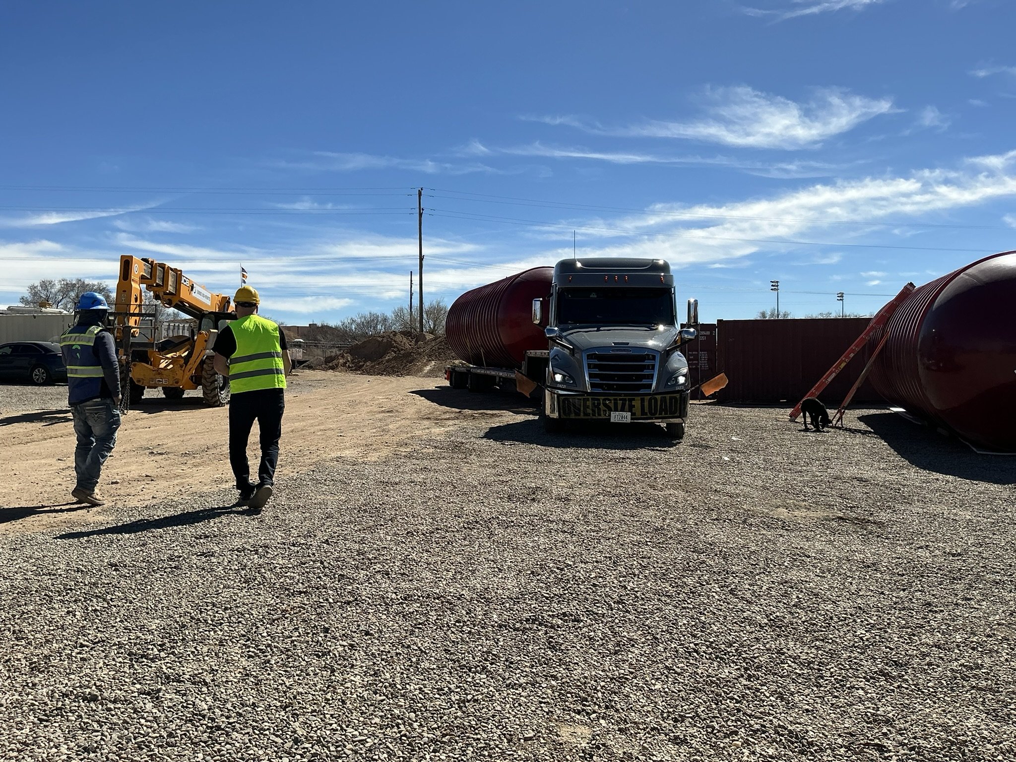 Construction site with two workers wearing safety vests and helmets, a large truck labeled 'Over Size Load,' a yellow construction crane, and two sizable red tanks stored upright on gravel ground under a blue sky with some clouds.