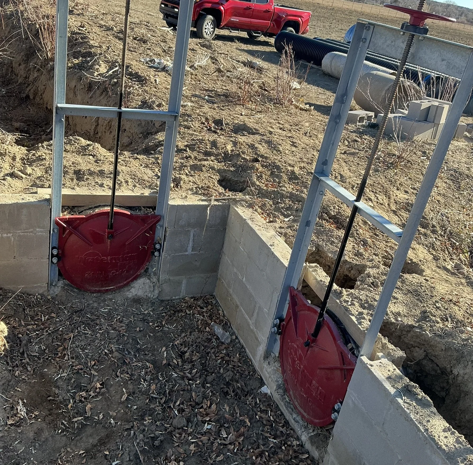 Two red fire hydrant access points with metal frames in an outdoor construction site, surrounded by dirt, gravel, and construction materials, with a red truck and pipes in the background.