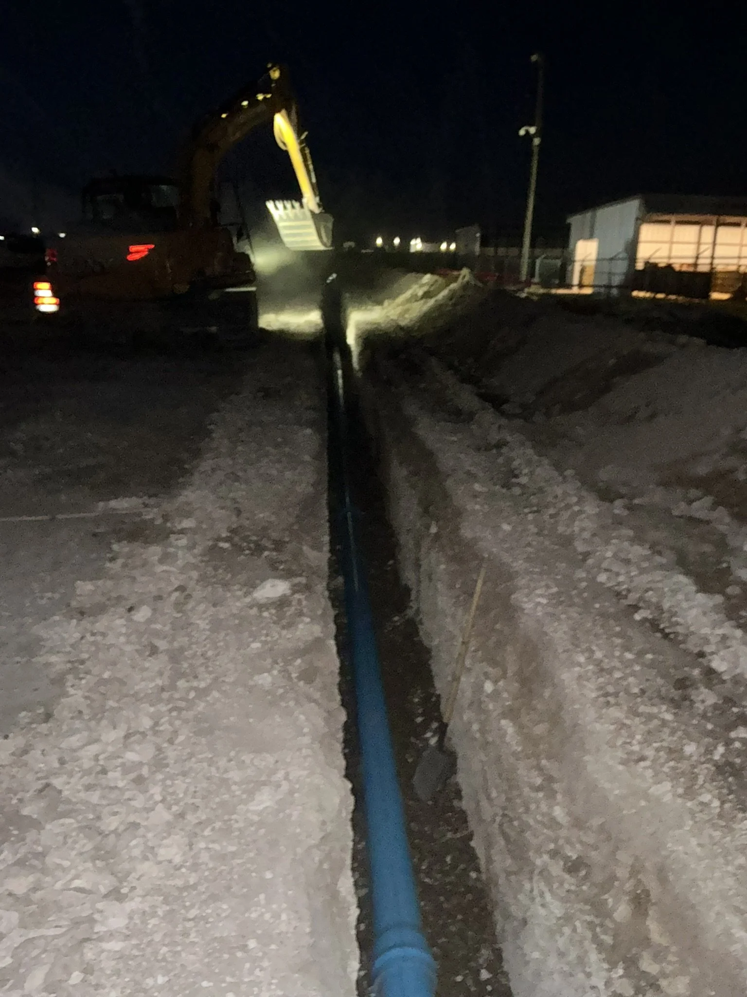 Nighttime scene with construction equipment, yellow excavator digging a trench, blue pipe laid in the trench, and a worker standing nearby. There is a building with a lit window in the background.