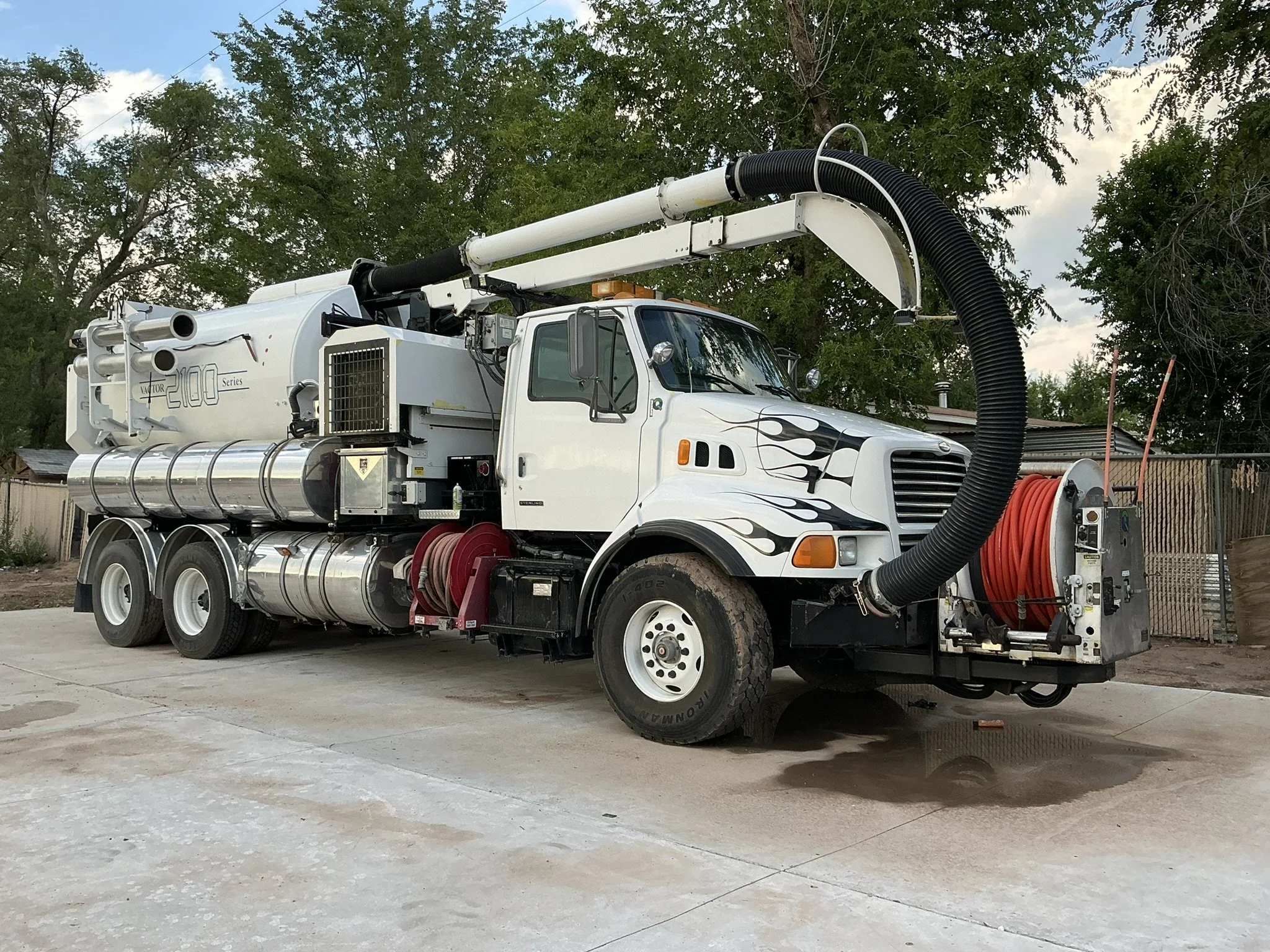 A large white vacuum truck with black and flame decals on the side, parked on a concrete surface in a backyard with trees and a fence in the background. The truck has a large hose connected to the front and a reel with orange hose on the side.