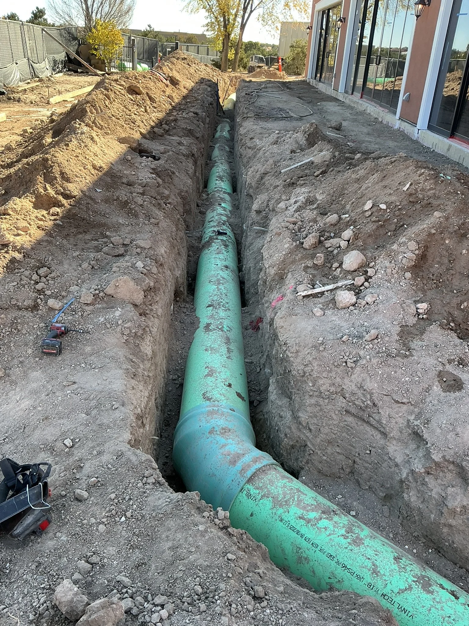 A construction site with a long trench containing a green pipe being installed, with dirt and rocks on the sides and a building with glass windows to the right.