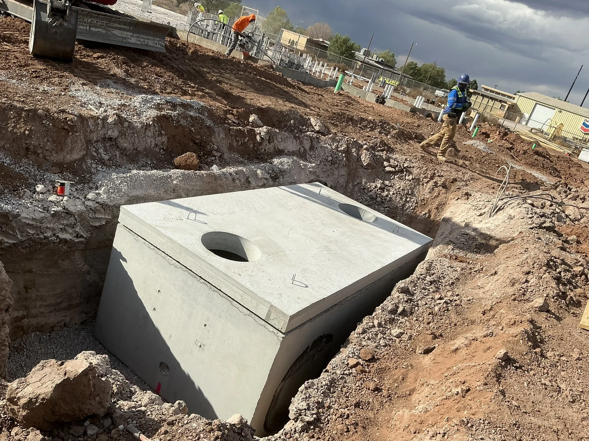 Construction workers are installing a large underground utility vault or septic tank in a dirt excavation site, with some workers in the background and cloudy sky overhead.