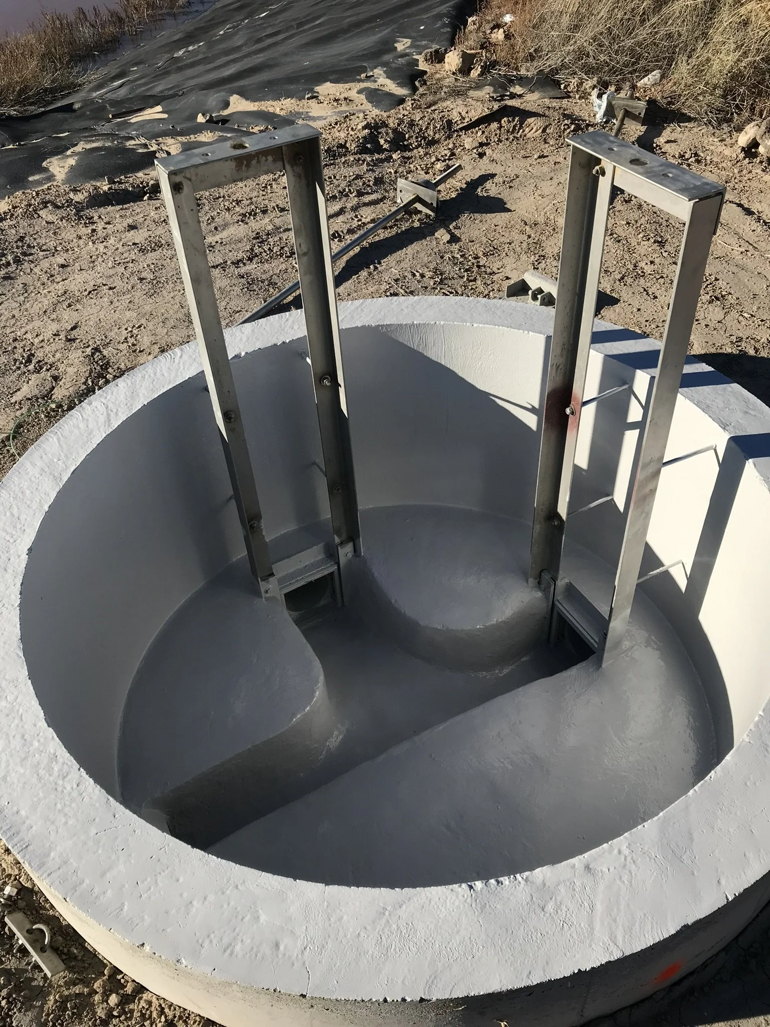 A concrete manhole with metal ladder rungs inside, situated outdoors on dirt ground with some rocks and bushes in the background.