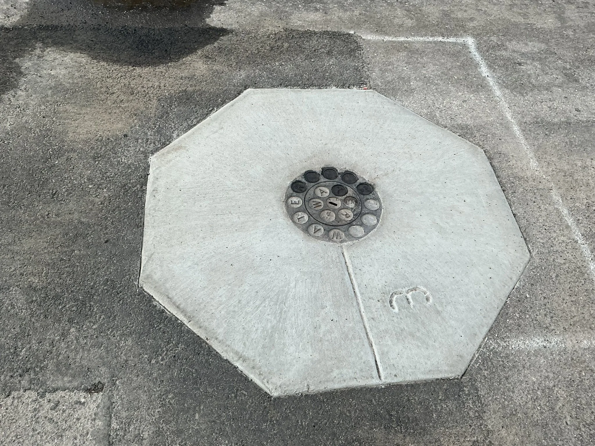 A white octagonal concrete marker on the ground with a circular metal plate in the center labeled with numbers and letters, and the number 3 inscribed on the concrete.