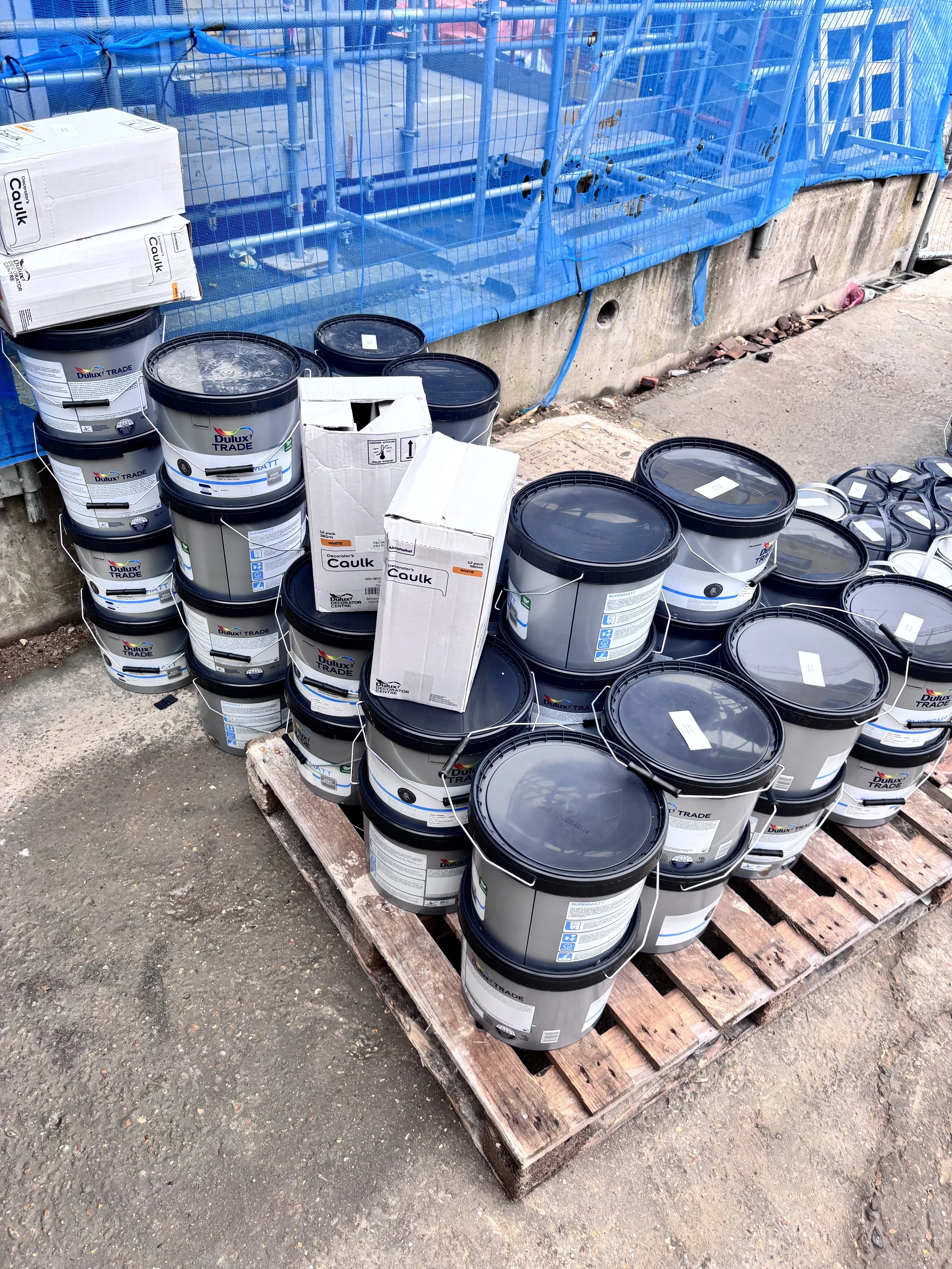 Stack of gray and white paint buckets labeled 'Dulux Trade' on a wooden pallet, with some boxes and blue netting in the background at a construction site.