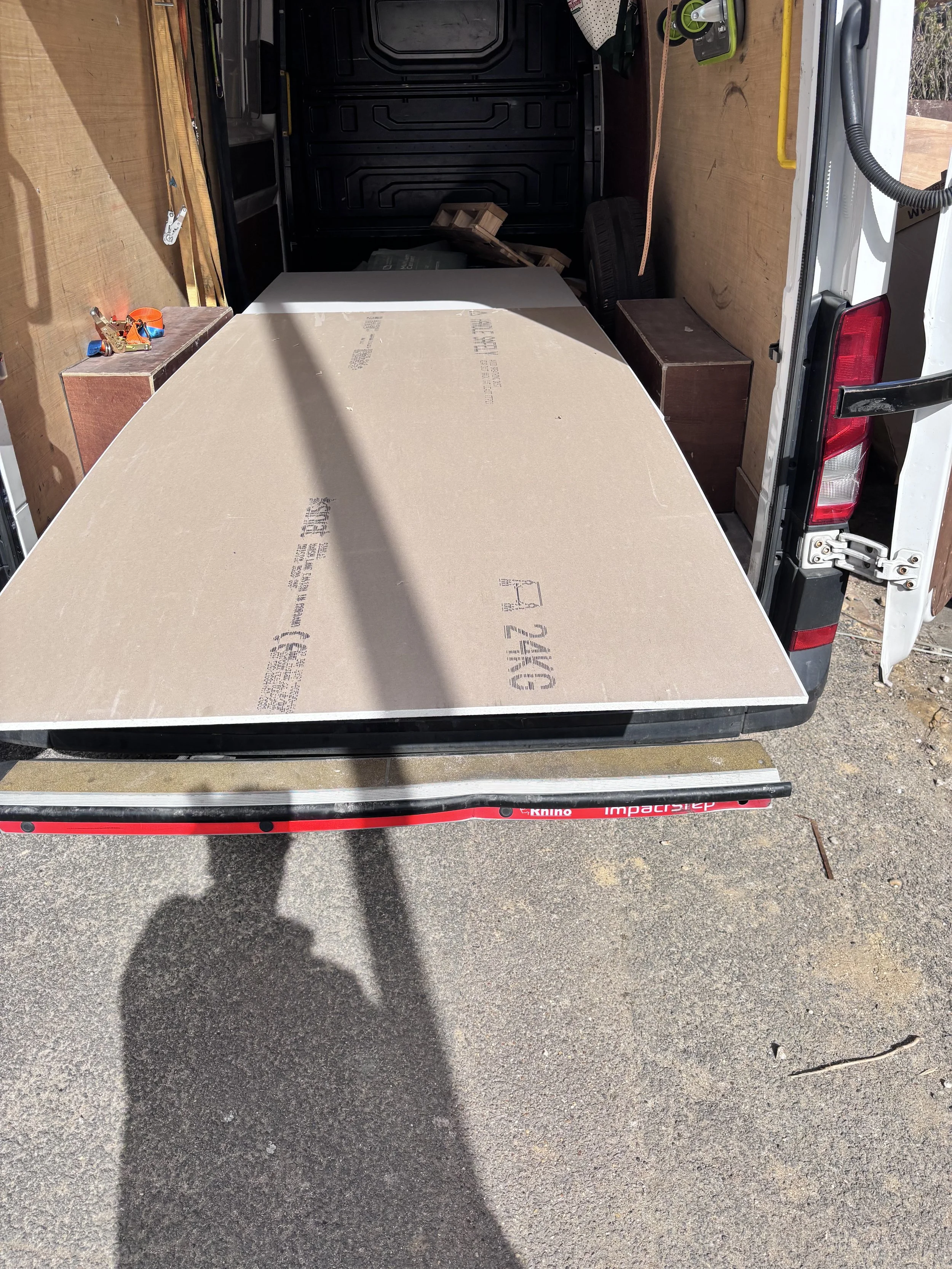 Open cargo van with wooden walls and a large piece of particle board inside. Tools and small items are on a small table on the left, with a tape measure and a small toolbox visible. A car tire and a wooden block are also inside the van.