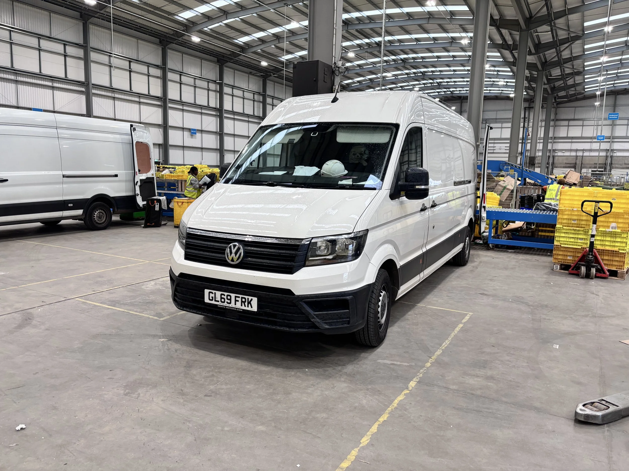 Interior of a warehouse with white Volkswagen van in foreground, workers in yellow vests, pallets, and equipment.