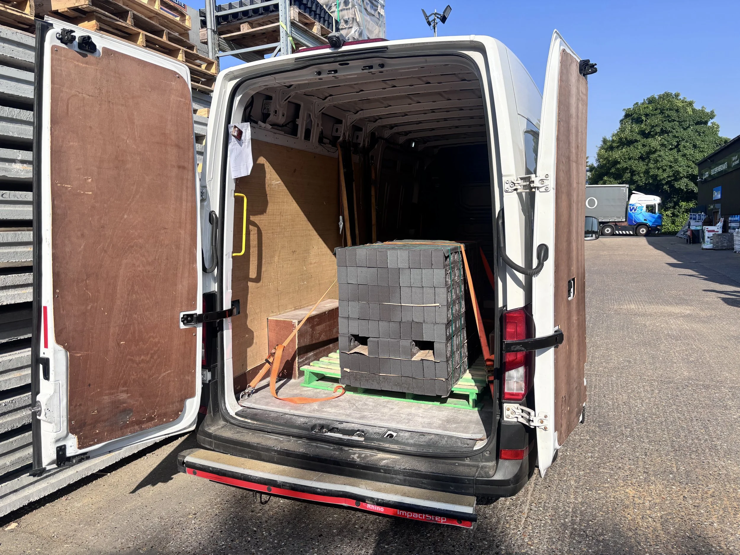 Open white delivery van with wooden panel interior, stacked with black paving stones, parked outdoors on asphalt near construction and storage area, under clear blue sky.