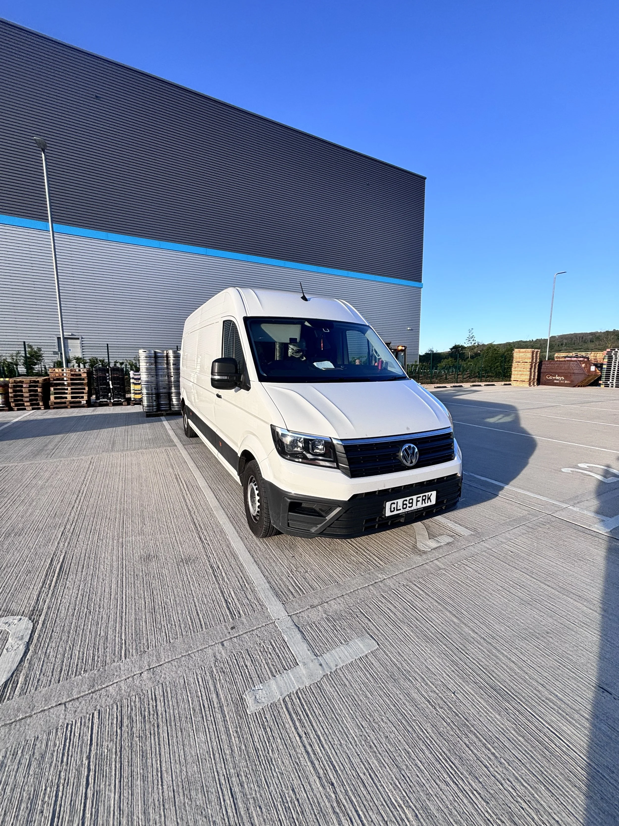White Volkswagen commercial van parked in an outdoor lot with pallets of wood and barrels behind it. Large industrial building with a blue stripe on the wall visible in the background.