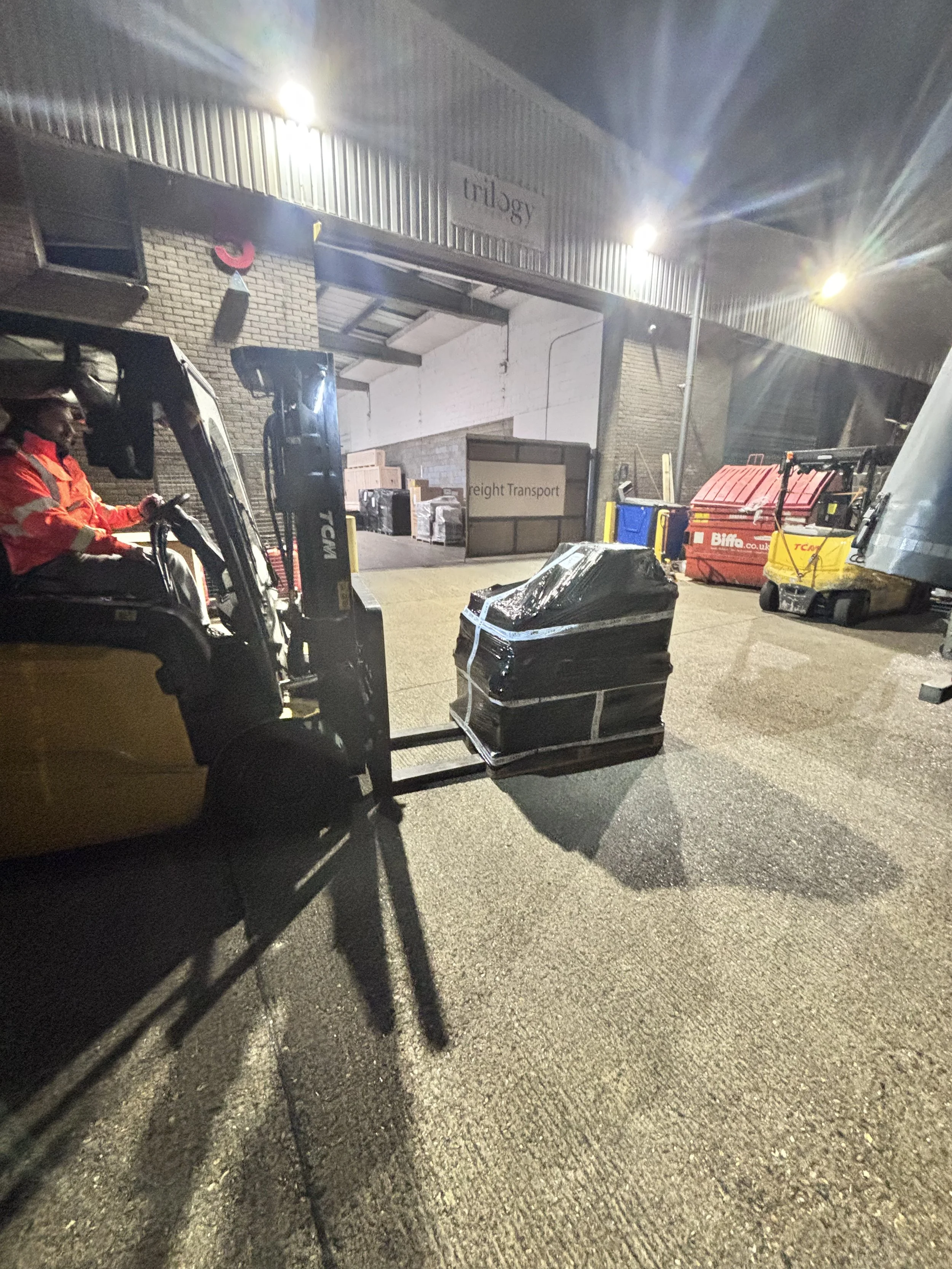 Nighttime scene outside a warehouse with a forklift, various trash bins, stacked boxes, and a person in an orange safety jacket operating the forklift. The warehouse has signs labeled 'trilogy' and 'freight transport', with bright lights illuminating
