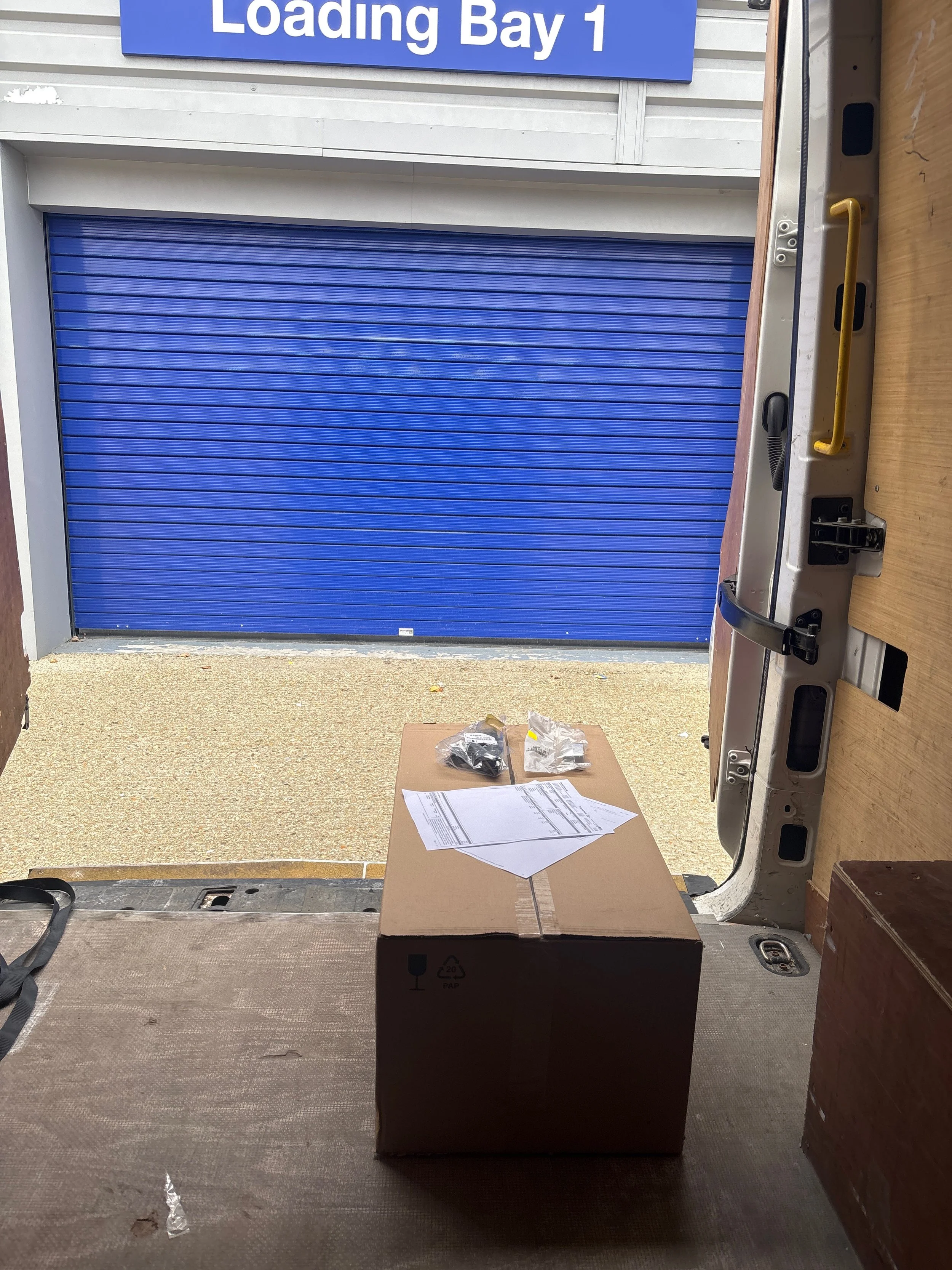 Loading Bay 1 garage door with a cardboard box, papers, and plastic bags on a table in the foreground, viewed from inside a delivery truck.