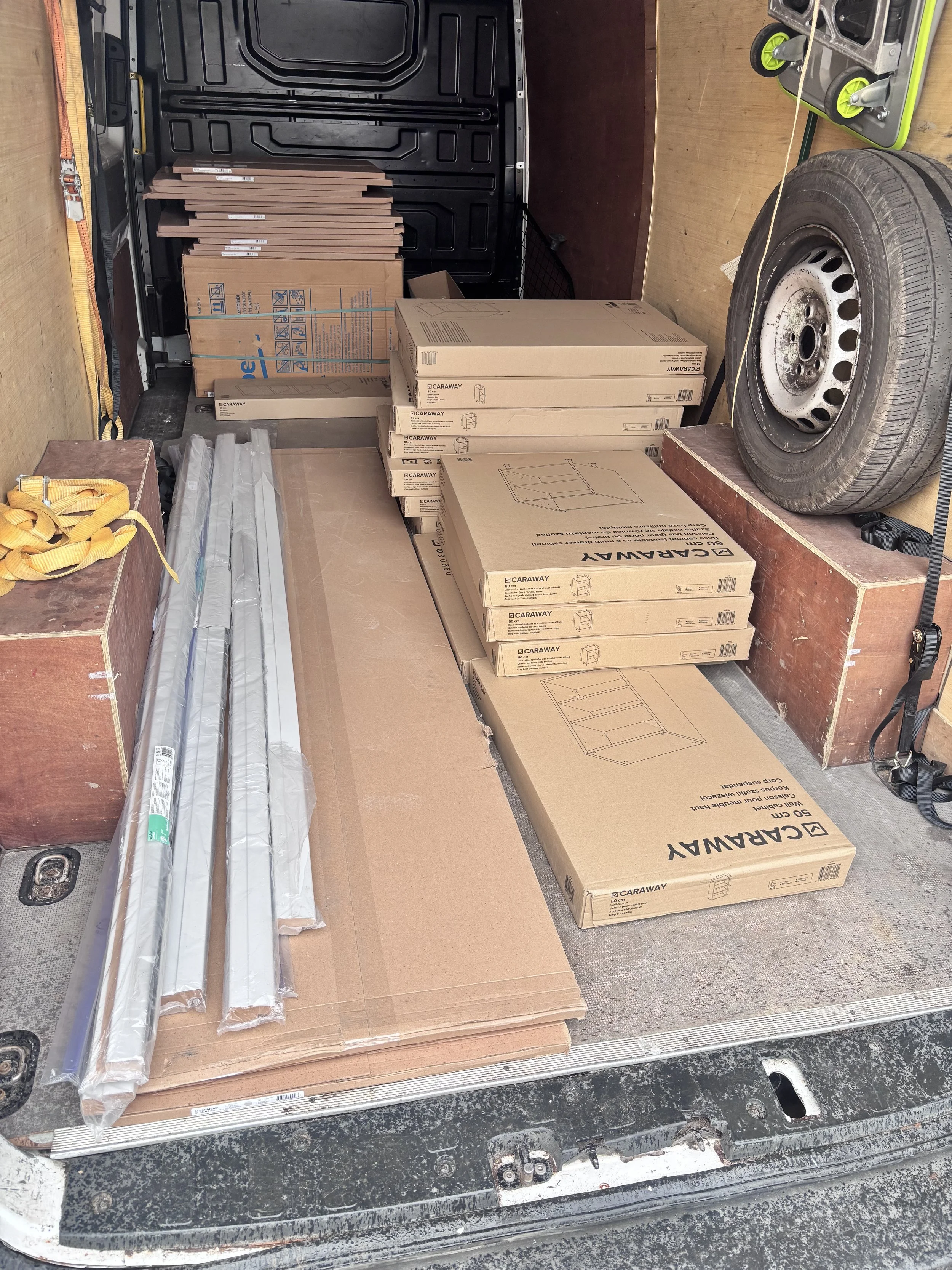 The image shows the open back of a vehicle filled with large cardboard boxes, some labeled 'Caraway' and others containing items like a tire and poles. Additionally, there is a small green dolly and yellow straps on the left.