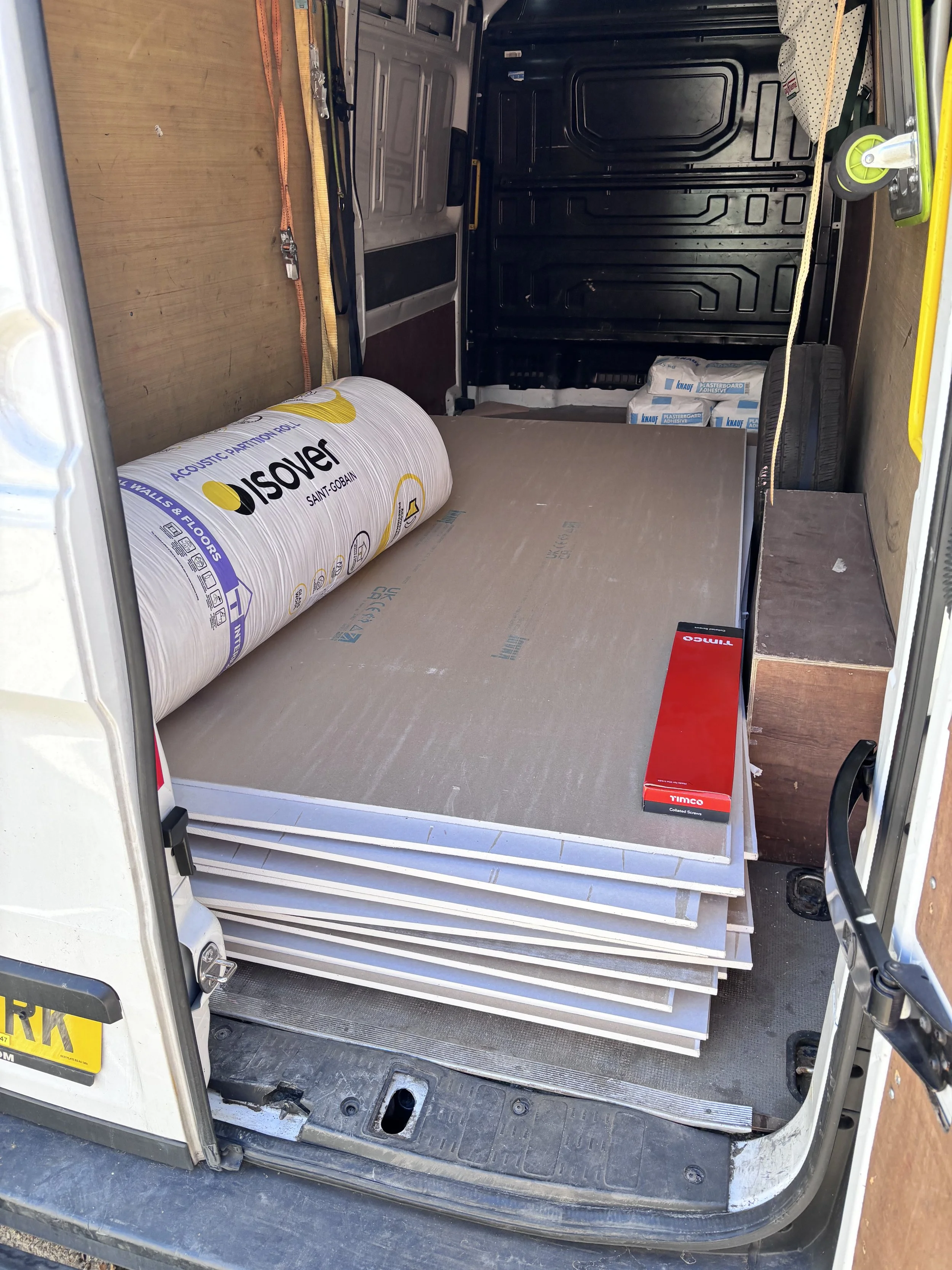 Inside of a van with a large roll of acoustic insulation material, some large sheets of drywall, and smaller boxes and tools stored inside.