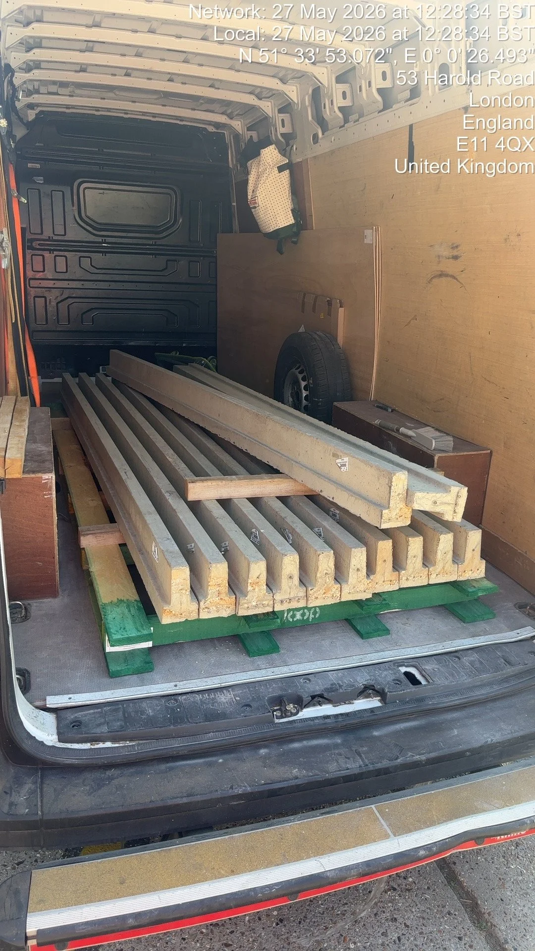 The back of a van filled with construction materials, including long concrete beams or pipes, wooden boards, a tire, and a small box. The interior of the van is lined with metal panels, and there are some tools and supplies visible.