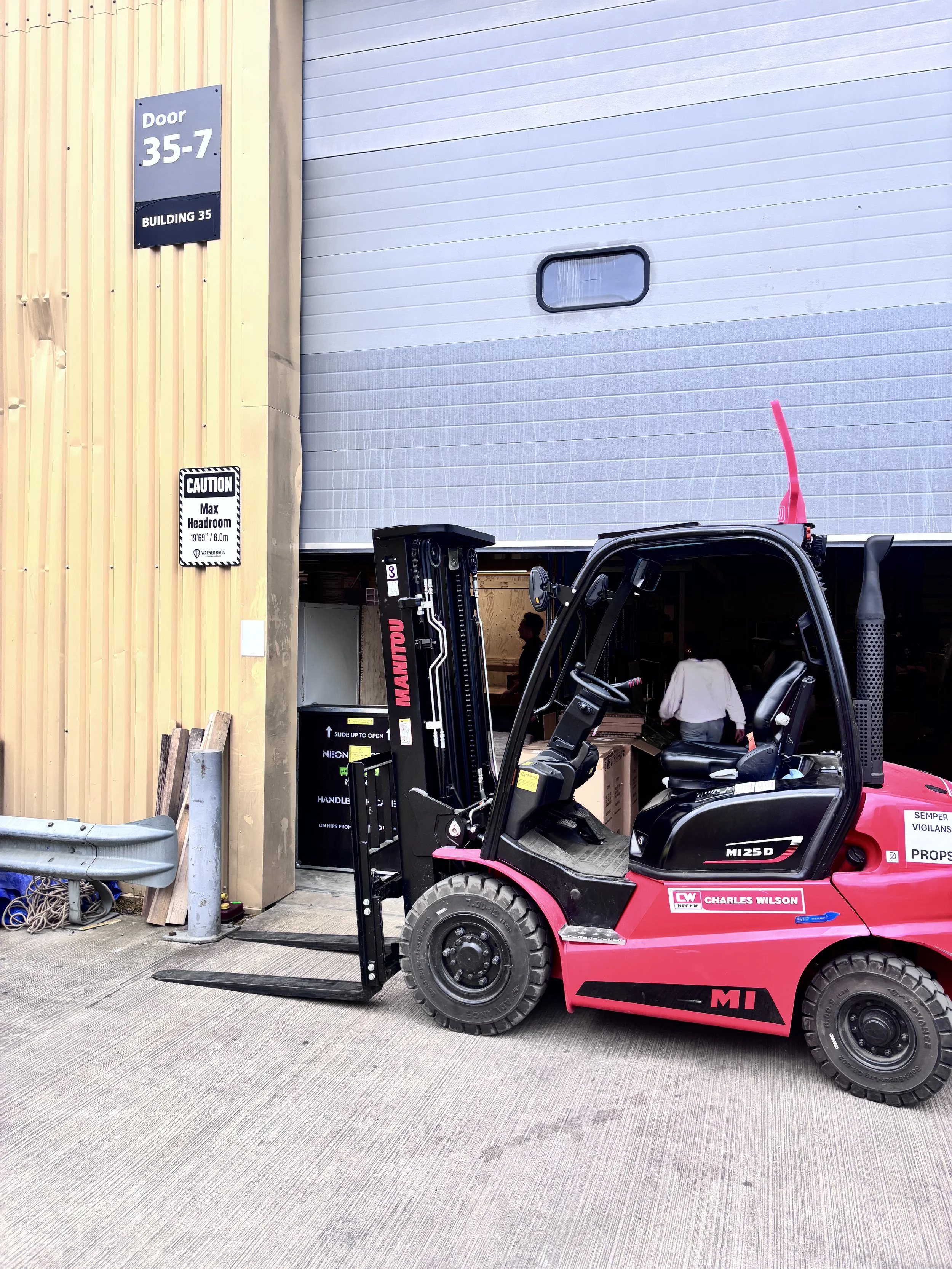 A red and black forklift parked outside a building with a partially open garage door. The building has a yellow vertical wood siding and a white metal garage door. There are two signs on the building: one indicating door number 35-7 and another cauti