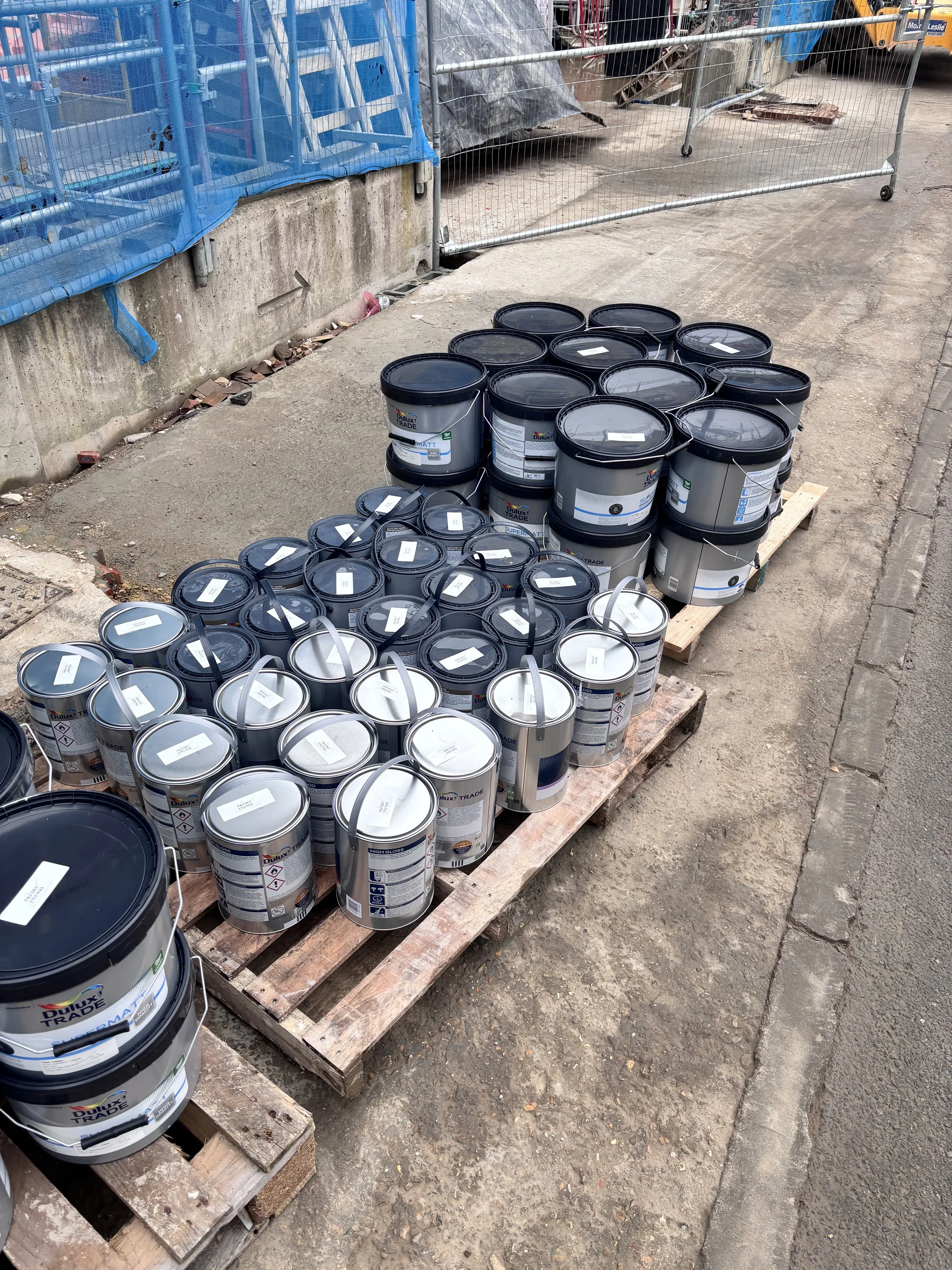 Multiple cans of Dulux Trade paint stacked on wooden pallets next to a construction site fence.