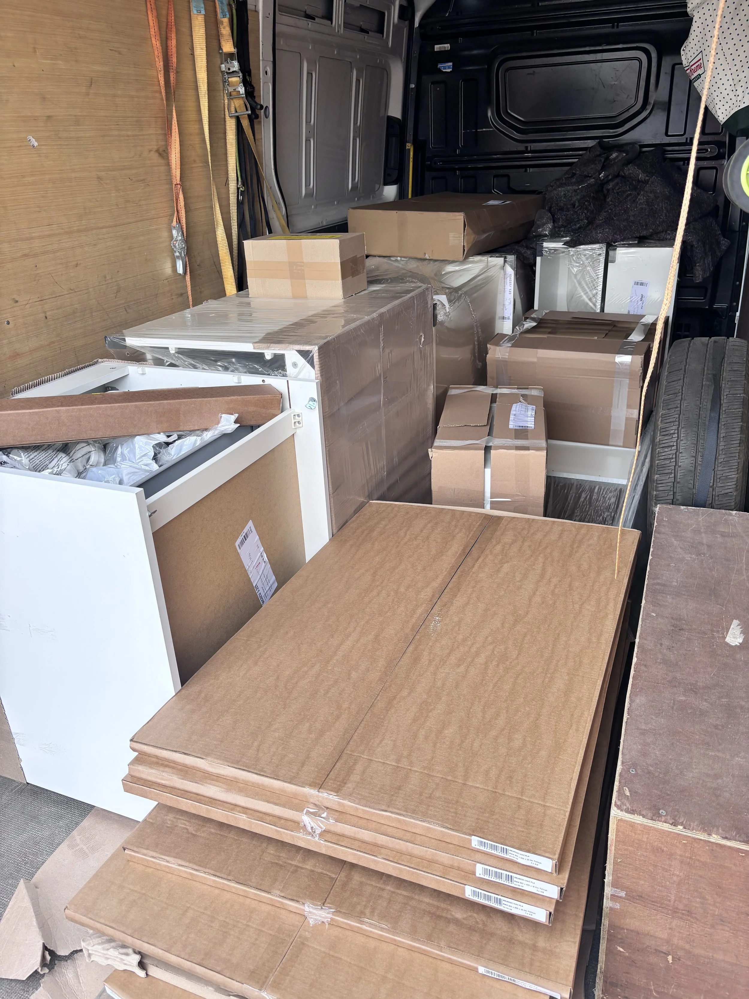 A store display with several boxed furniture pieces made of wood or wood-like material, arranged on a beige tiled floor. The boxes are wrapped in plastic, and some have shipping labels. The background features black walls with a yellow strip, part of