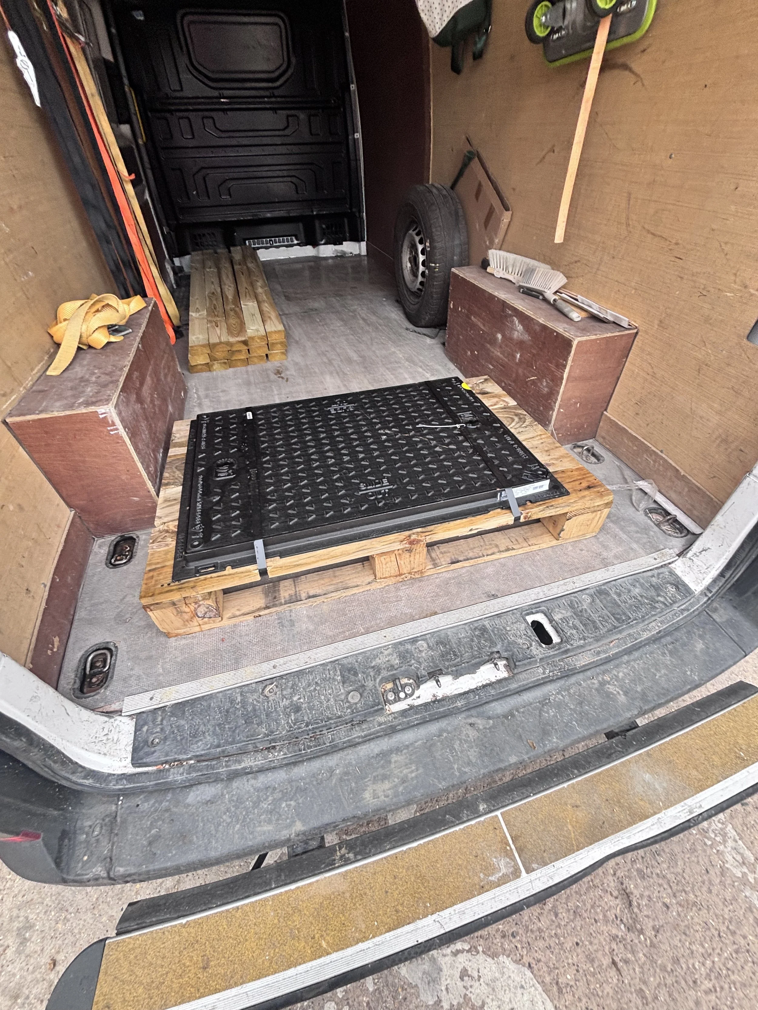The image shows the back of a van with the rear door open, revealing the interior. Inside, there is a stack of wooden planks, a spare tire, and various tools and items. The floor has a black grid panel, and the van's interior walls are wood-paneled.