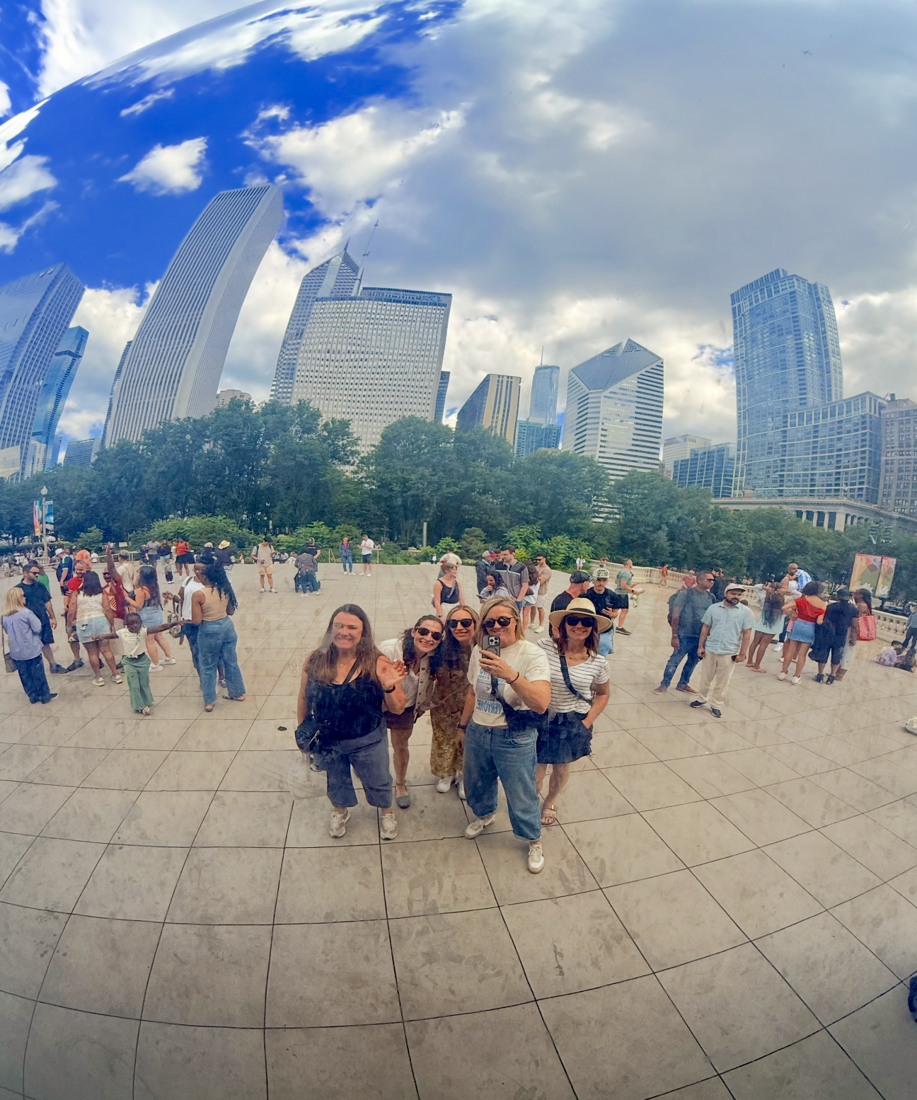 A group of friends taking a selfie in a busy city plaza surrounded by tall skyscrapers under a partly cloudy sky.
