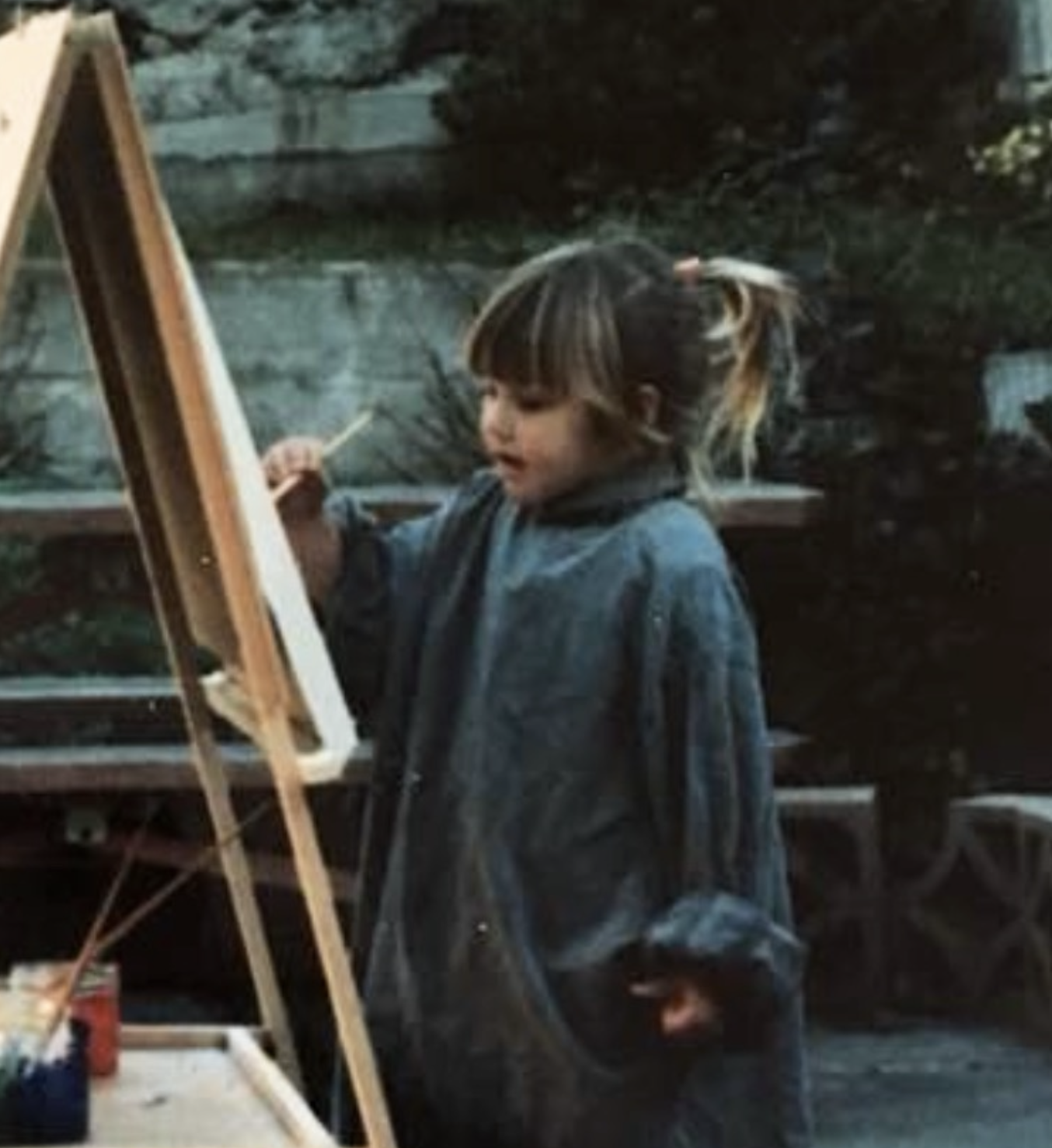 A young girl with a ponytail painting on an easel outdoors, with trees and a stone wall in the background.