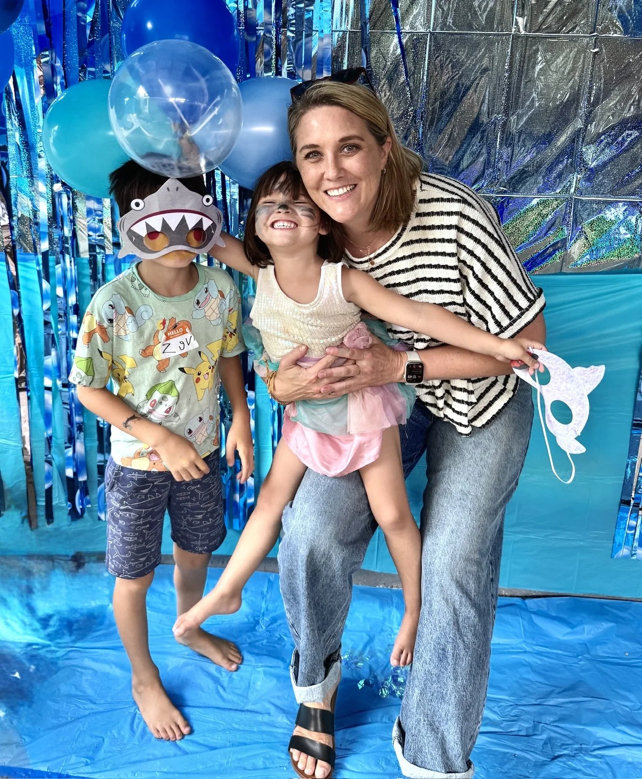 A woman with two children in a festive setting. One child wears a dinosaur mask, and the other is smiling with face paint. Blue balloons and shiny blue and silver decorations are in the background.