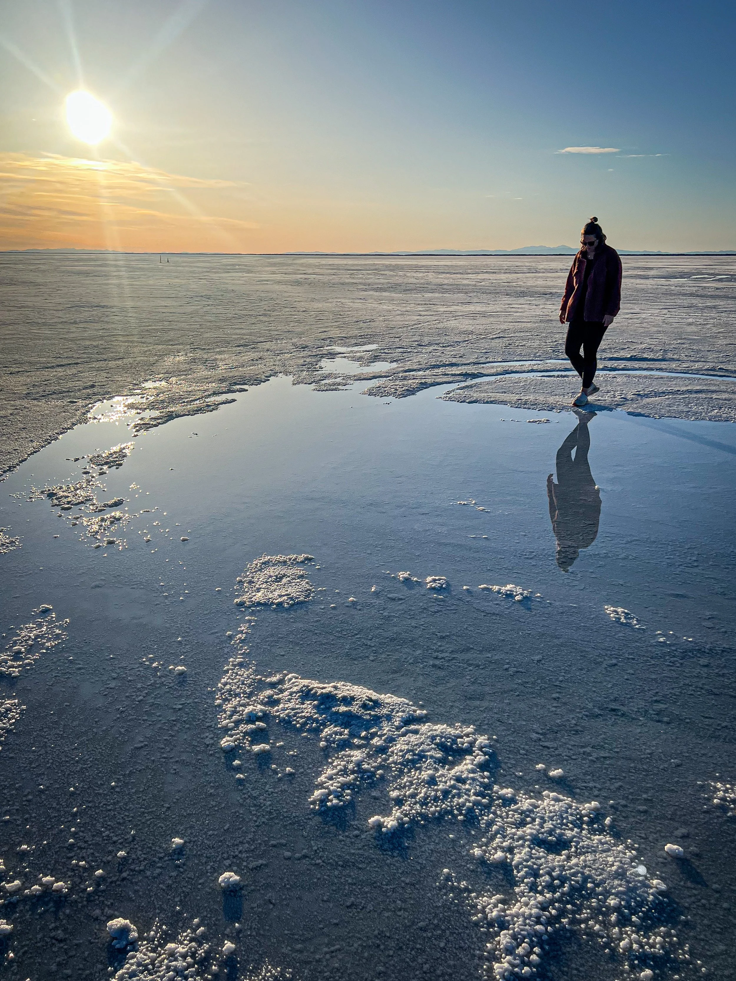 A person walking on a snow-covered landscape near a shallow pool, with the sun low in the sky and mountains in the distance.