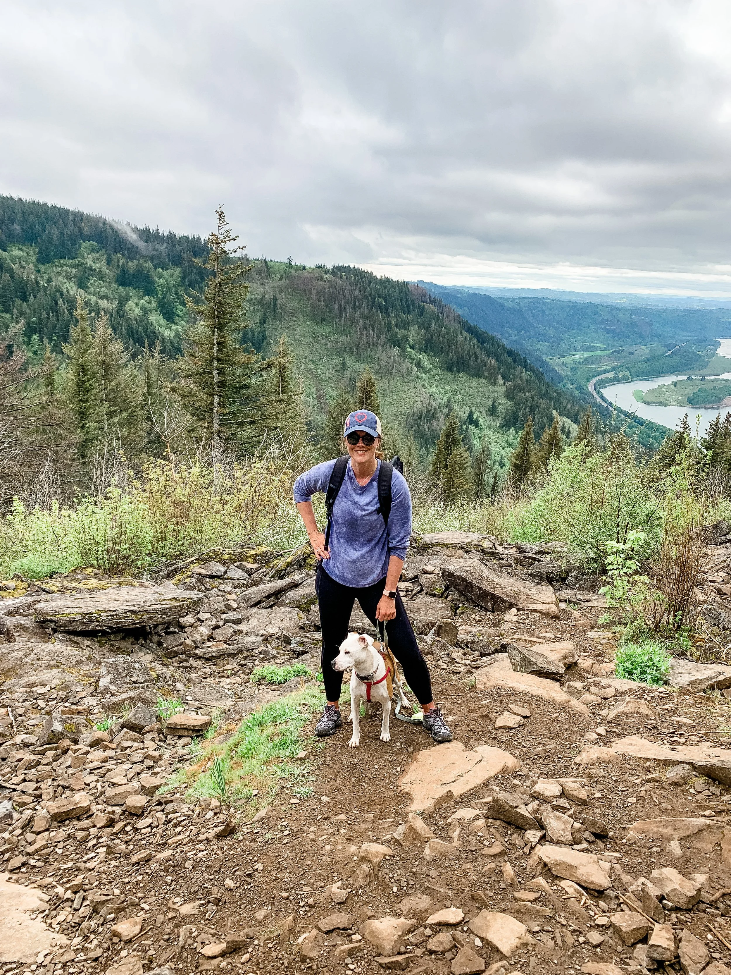 A person in gray athletic clothing and a gray hat smiling at the camera while standing on a rocky trail with a white dog on a red leash. They are in a mountainous area with forested slopes and a winding river in the background under a cloudy sky.
