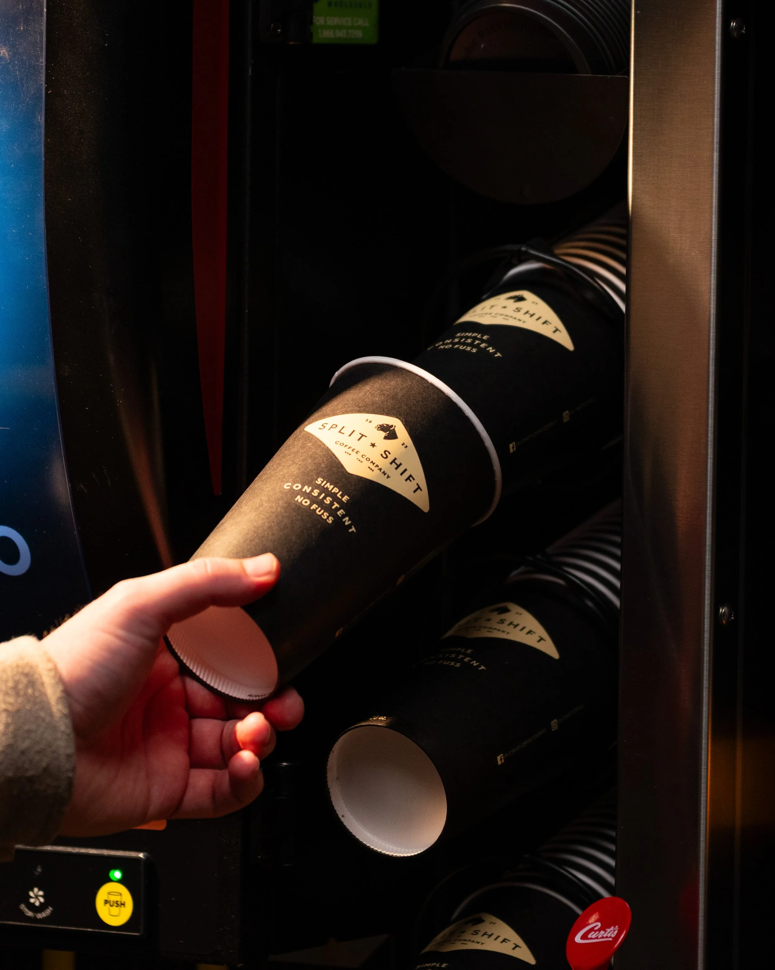 A person holding a black cup with a beige label that reads 'Split + Shift Coffee Company' and 'Simple, Consistent, No Fuss' in front of a coffee vending machine with multiple cups stored inside.