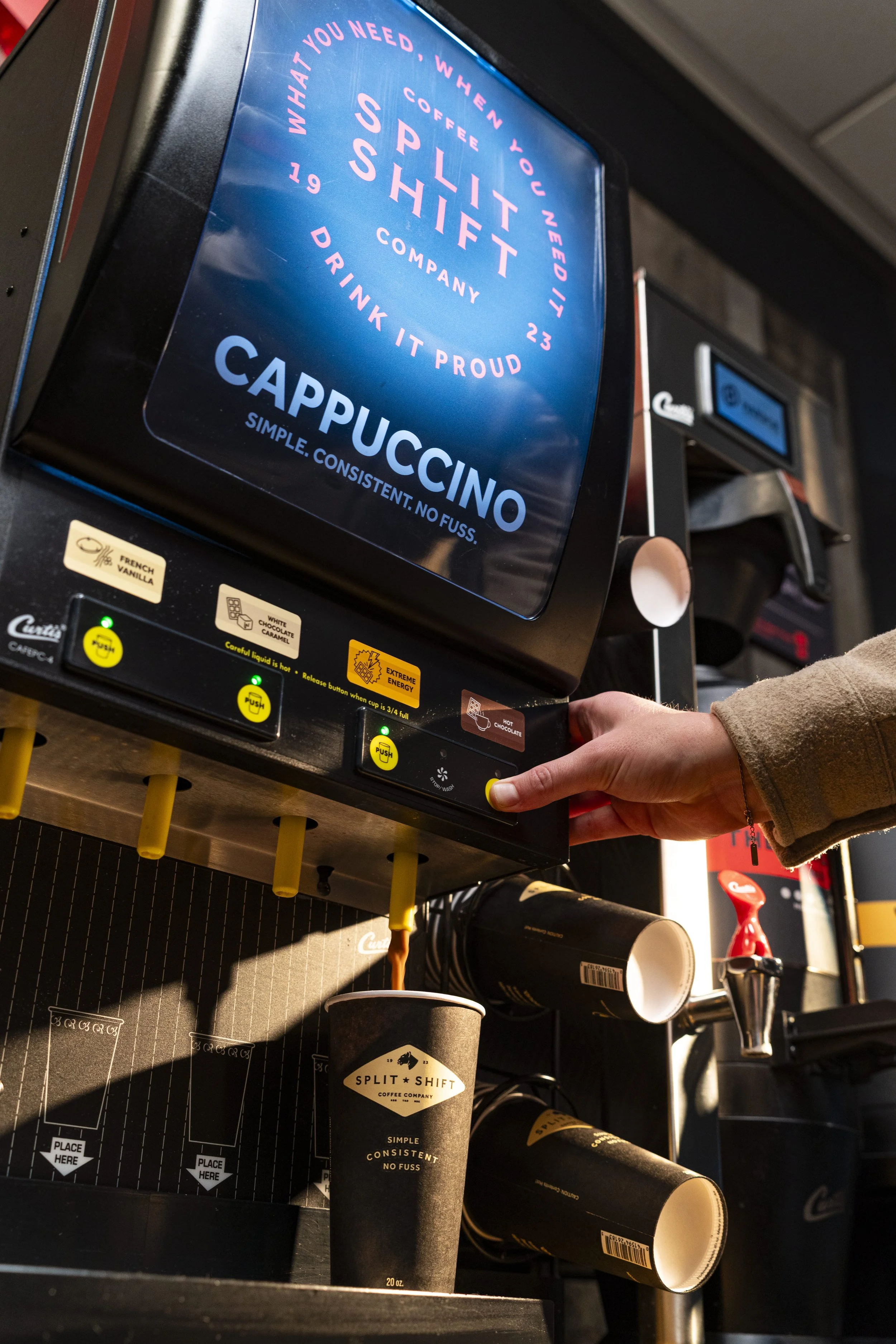 A digital menu display promoting coffee and drinks at a coffee shop. The menu highlights cappuccino with the slogan "Simple. Consistent. No Fuss." and other beverages like split shift coffee. The display is mounted above the counter with disposable cups and coffee equipment visible below.