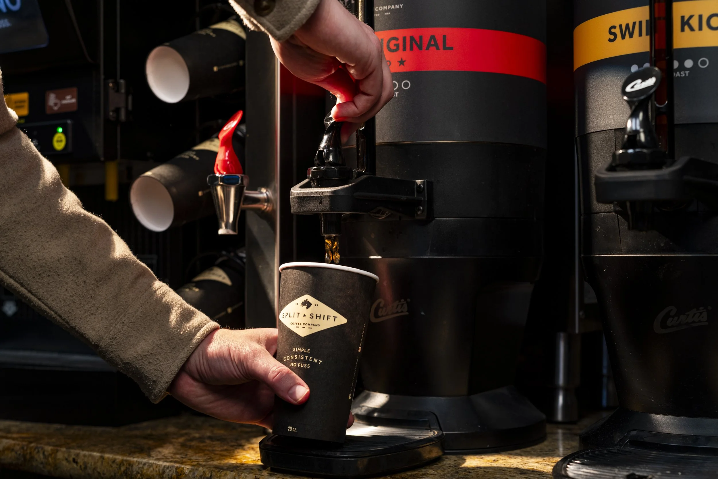 A person's hand, wearing a beige sweater sleeve, is holding a black paper coffee cup with a black lid. The cup has a logo that reads 'Split Shift Coffee Company.' The person is pouring coffee from a coffee machine with a red and black label into the cup. In the background, part of the coffee machine is visible with other cups stacked, labeled 'Original' and 'Swit Kick.' The setting appears to be a coffee shop or café.