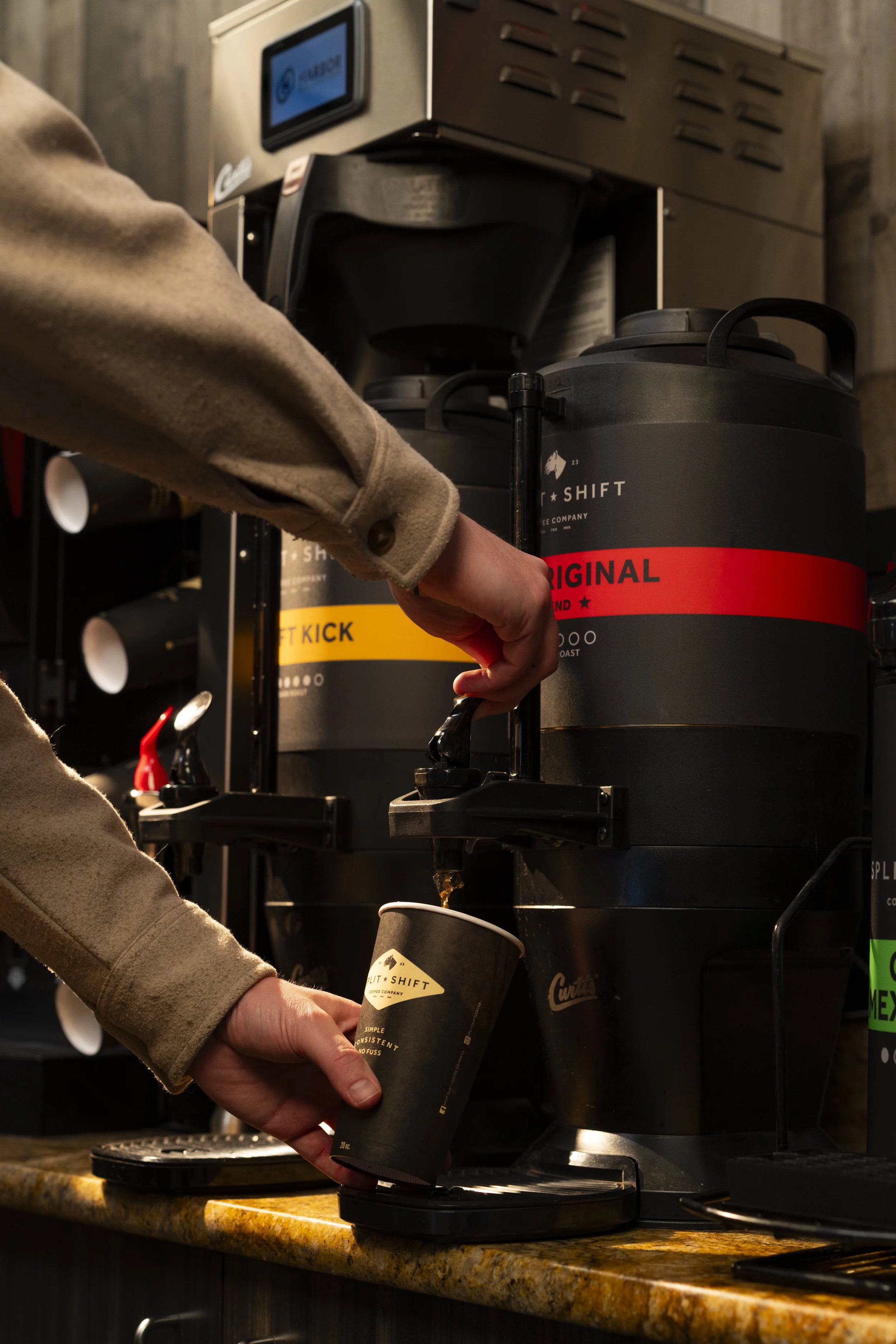 Person wearing a tan coat filling a black paper coffee cup from a black coffee dispenser machine in a coffee shop.