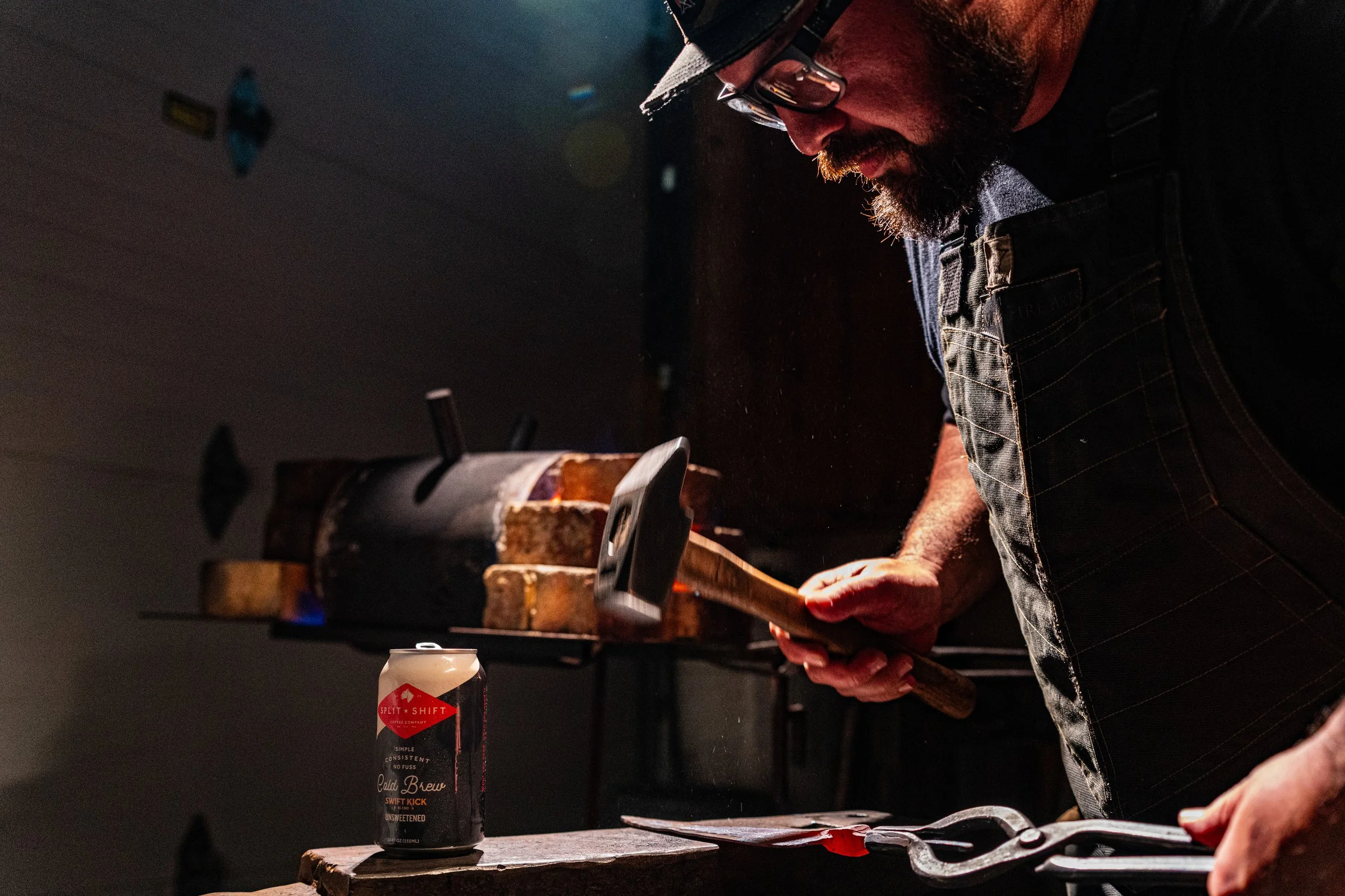 A man with a beard and glasses wearing a cowboy hat and plaid shirt working on a woodworking project, using a hammer on a piece of wood with a vise in a dark workshop. There is a can of cold brew coffee on the table.