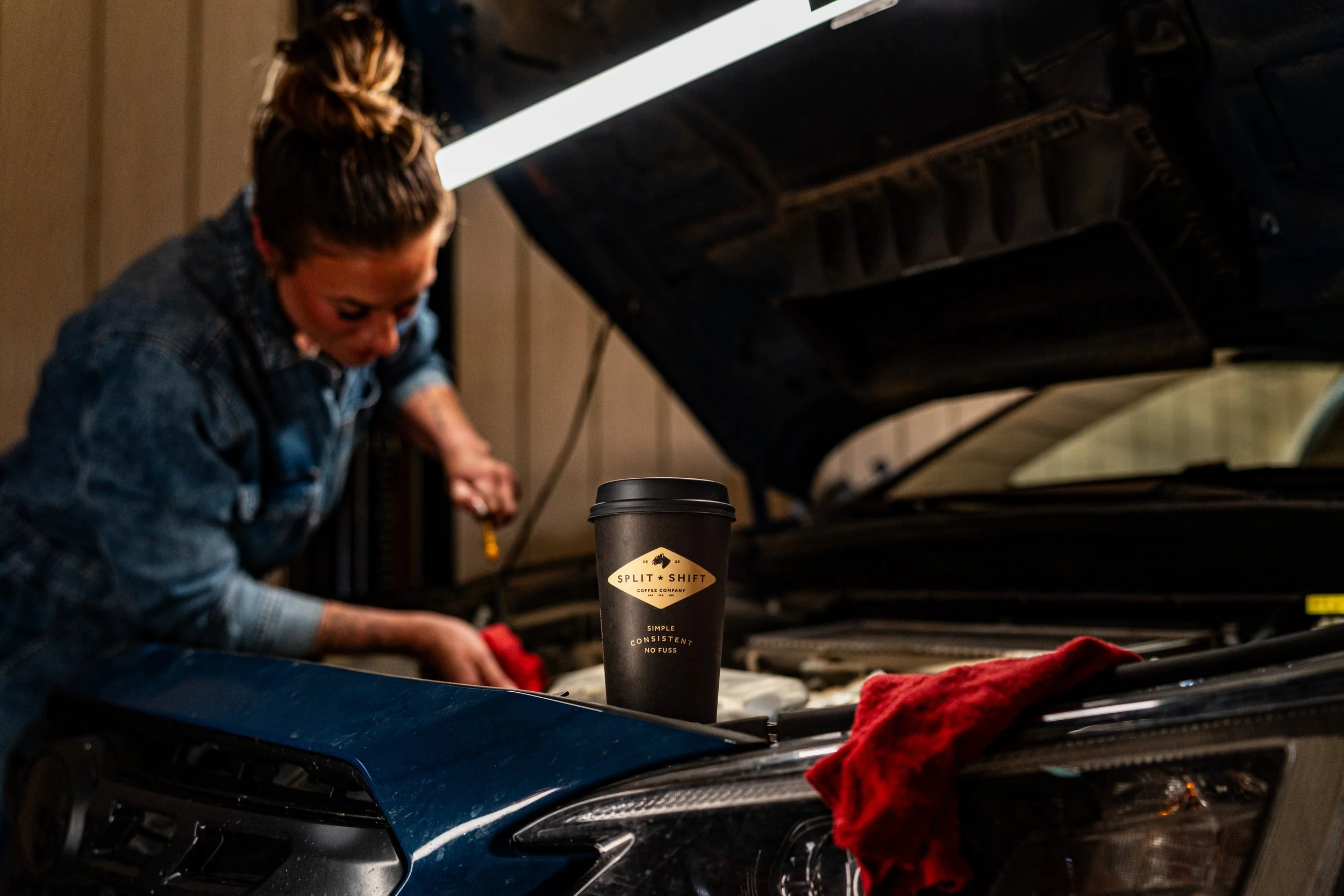 A woman is working under the hood of a blue car, with a coffee cup in the foreground that has the logo 'Split Shift Coffee Company.'