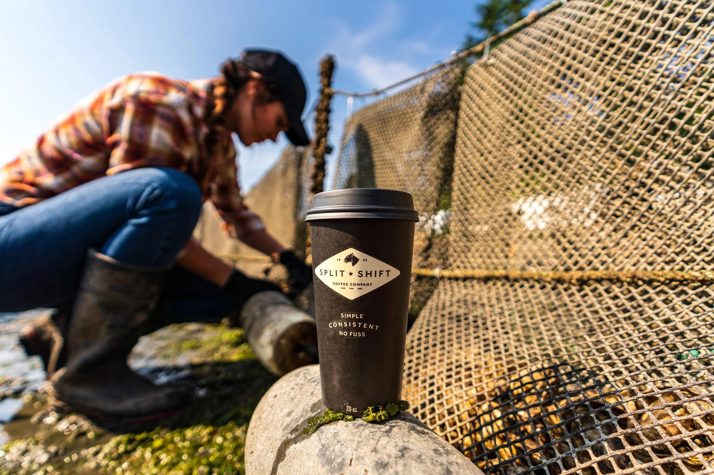 A person crouching near a fence, wearing a plaid shirt and a cap, with a coffee cup in the foreground on a rock. The coffee cup has a label with a logo and text, outdoor setting with blue sky.