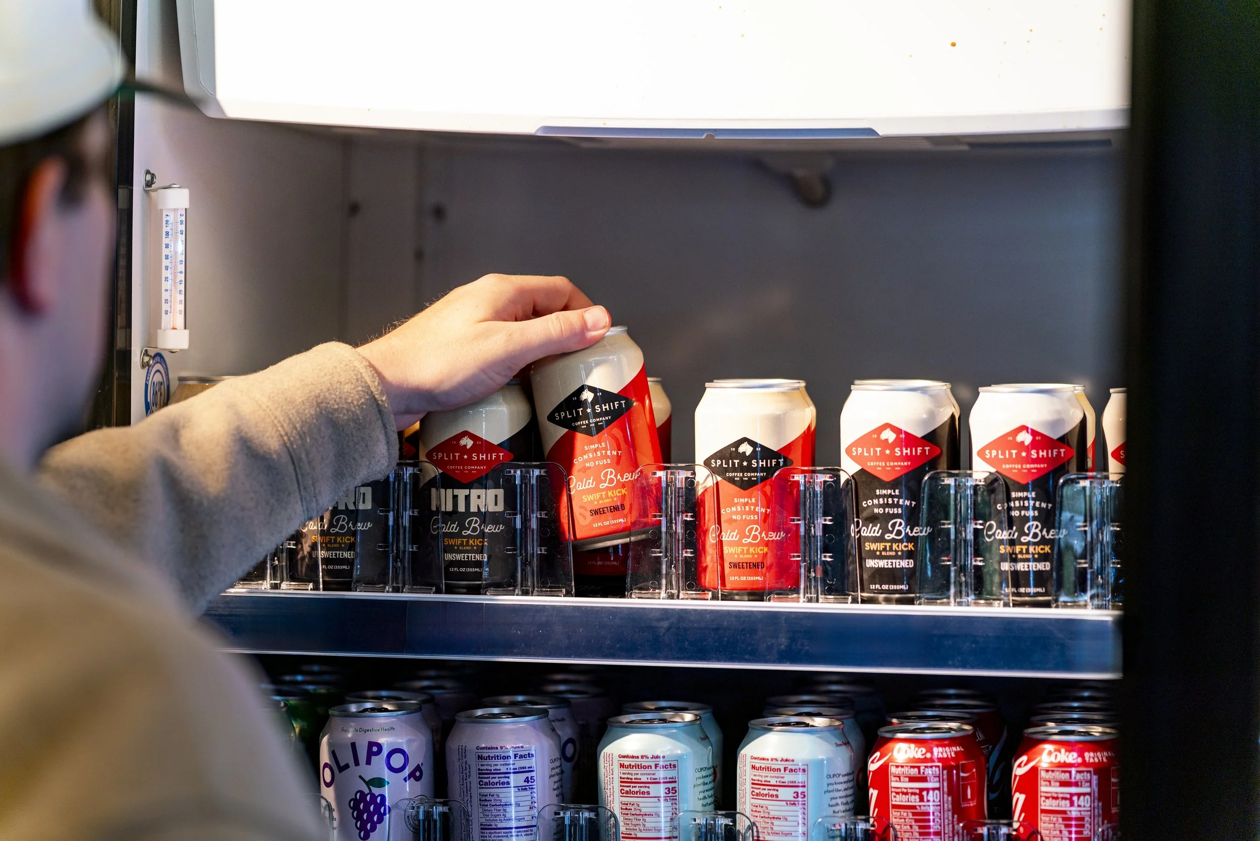 A person is reaching into a refrigerator to grab a can of cold coffee. The refrigerator contains various canned drinks, including a variety of soda and flavored beverages.
