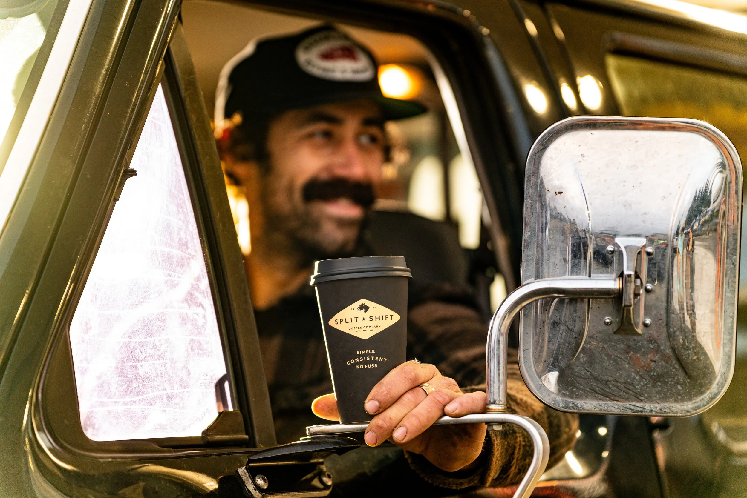 A man with a beard and mustache, wearing a black cap, is handing a cup of coffee through a car window. The coffee cup has a label from Split Shift Coffee Company. The man is smiling and the background is outdoors.