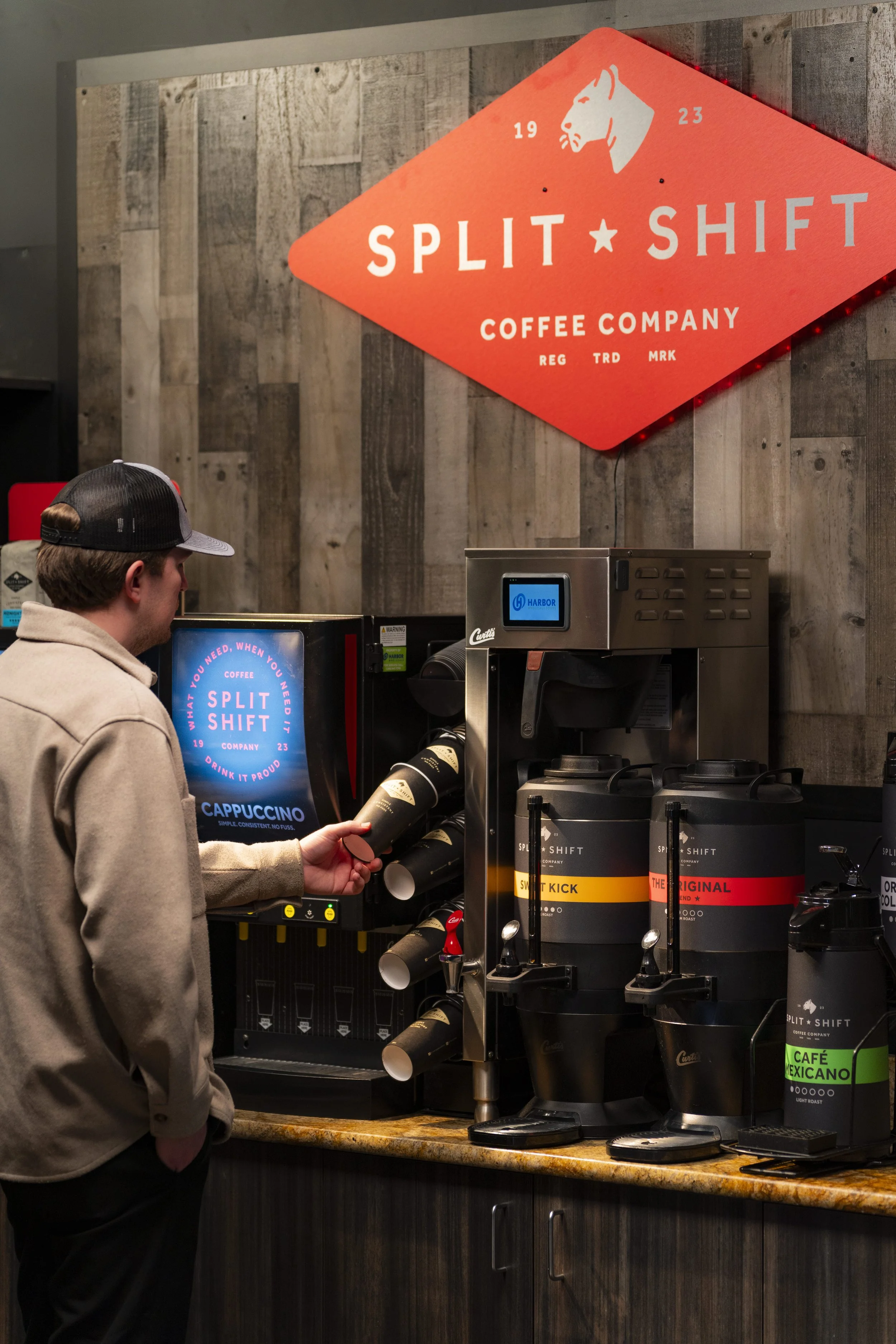 A young man at a coffee shop counter, filling his cup from a coffee dispenser labeled 'Split Shift Coffee Company' with various coffee options, in front of a wooden wall with a red sign displaying the company logo and name.
