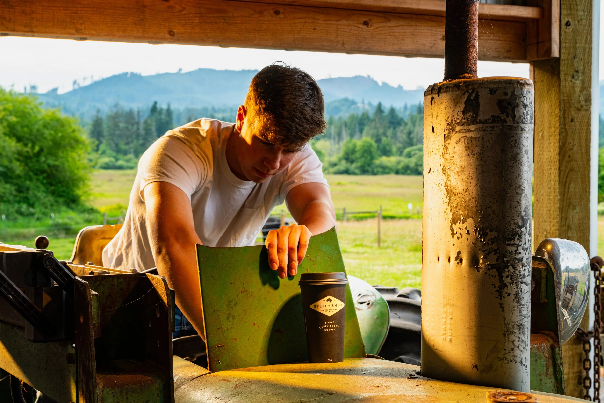 A young man with dark hair working outdoors among machinery and equipment, with a scenic view of green trees and mountains in the background at sunset.